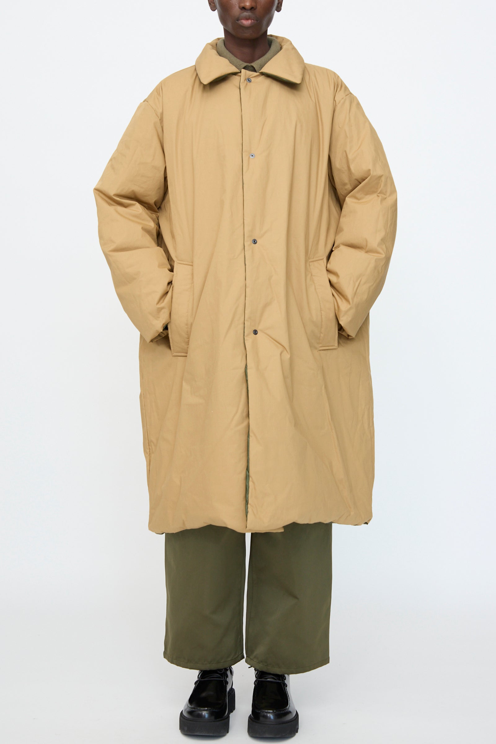 A person wears the Sofie D'Hoore Reversible Otis Down Coat in Mushroom and Khaki over olive green pants and black shoes, standing against a plain white background.