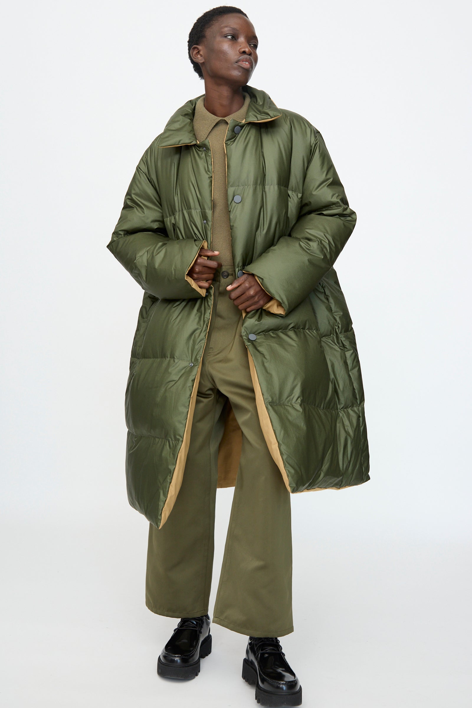 A model wears the Sofie D'Hoore Reversible Otis Down Coat in Mushroom and Khaki over a green outfit with black platform shoes, posed against a plain white background.