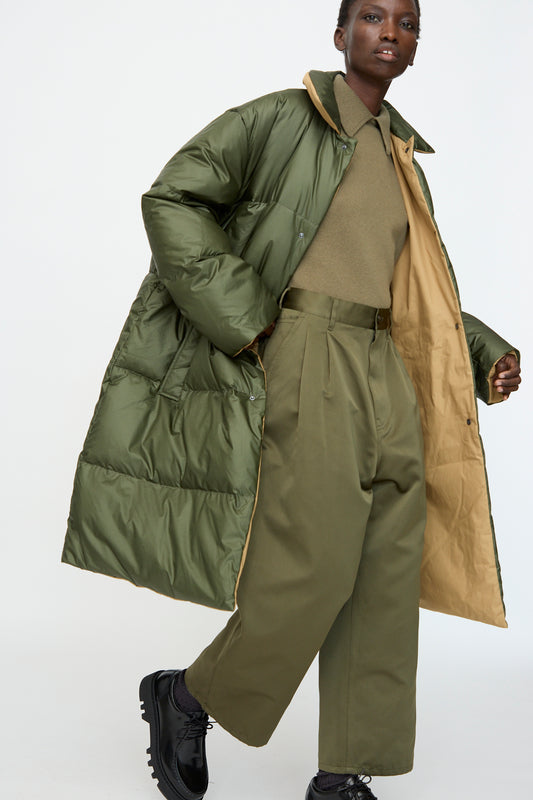 A person wears the Sofie D'Hoore Reversible Otis Down Coat in Mushroom and Khaki, paired with wide-leg high-waisted trousers, a matching sweater, and black shoes against a plain background.