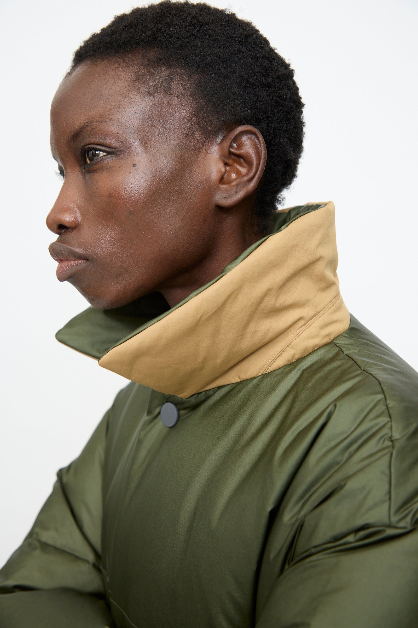 Person in profile wears the Sofie D'Hoore Reversible Otis Down Coat in Mushroom and Khaki, featuring a high tan collar, looking left against a plain white background.