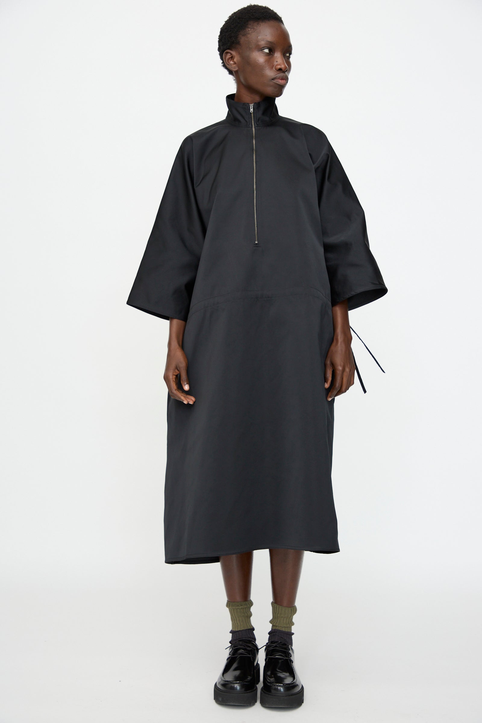 A person in the Sofie D'Hoore Satin Dolci Dress in Black—a loose-fitting, long midi dress with a high collar, half-zip front, and wide sleeves—pairs it with olive socks and black shoes against a plain white background.