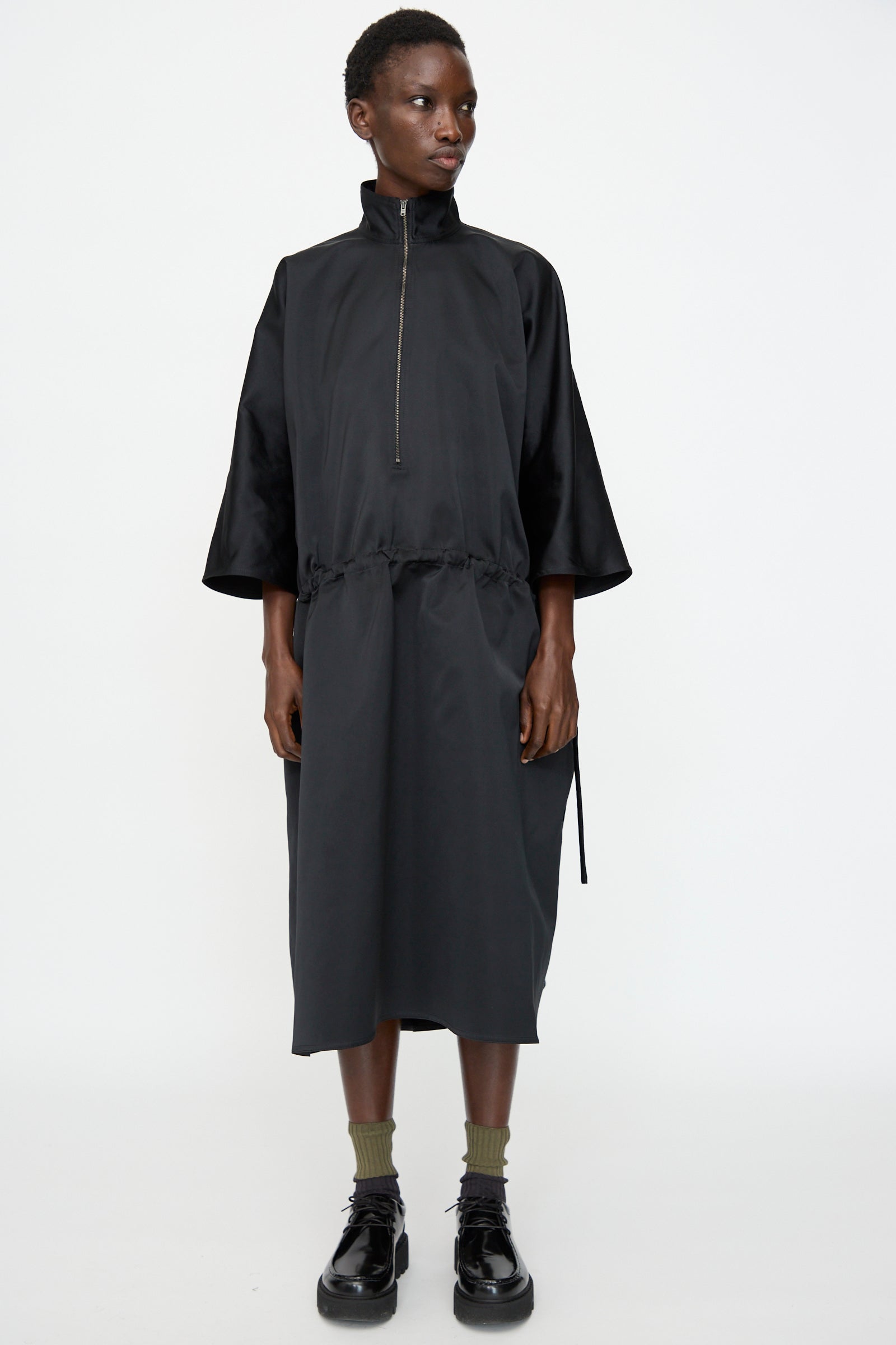 A person stands against a plain white background wearing the Sofie D'Hoore Satin Dolci Dress in Black, featuring long sleeves, a zip neck, drawstring waist, and crafted from satin cuir polyester. Styled with green socks and shiny black shoes.