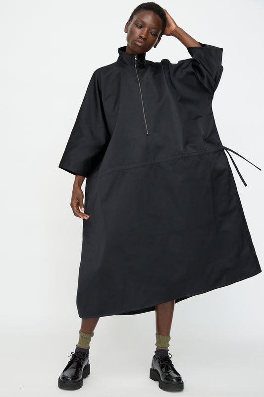 Wearing Sofie D'Hoore's Satin Dolci Dress in Black—a loose, mid-length satin cuir polyester piece with front zipper and tie detail—paired with black platform shoes and olive green socks.