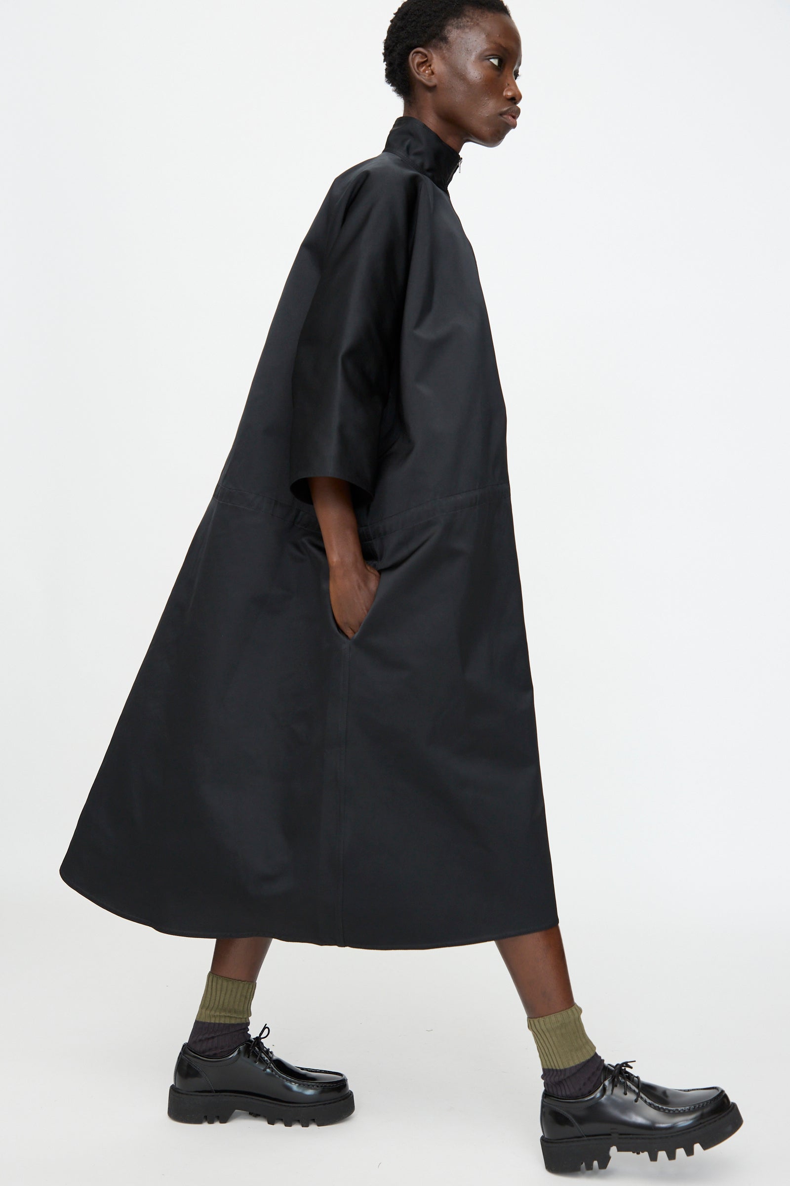 A person wears the Sofie D'Hoore Satin Dolci Dress in Black—a loose, half-sleeve midi dress in satin cuir polyester—paired with green socks and black chunky shoes, walking against a plain white background.