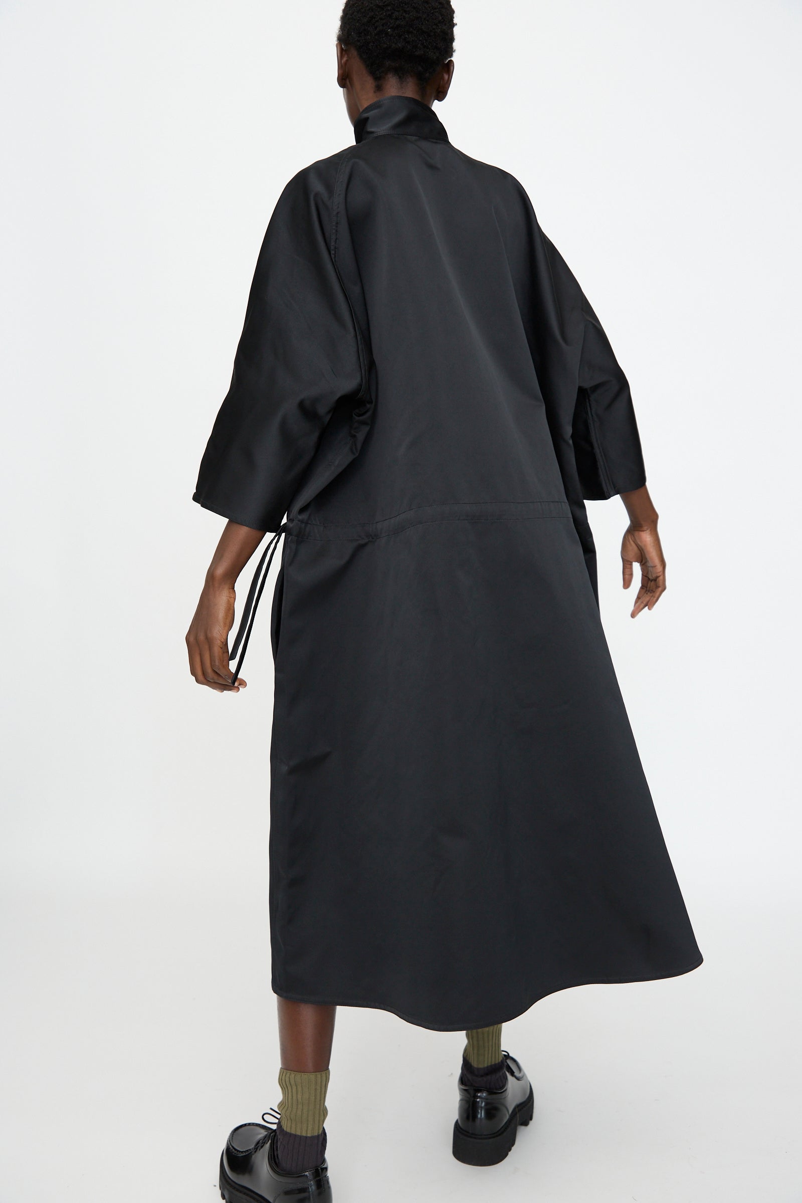 Model wears the Satin Dolci Dress in Black by Sofie D'Hoore, styled with olive green socks and black platform shoes, standing against a plain white background and facing away from the camera.