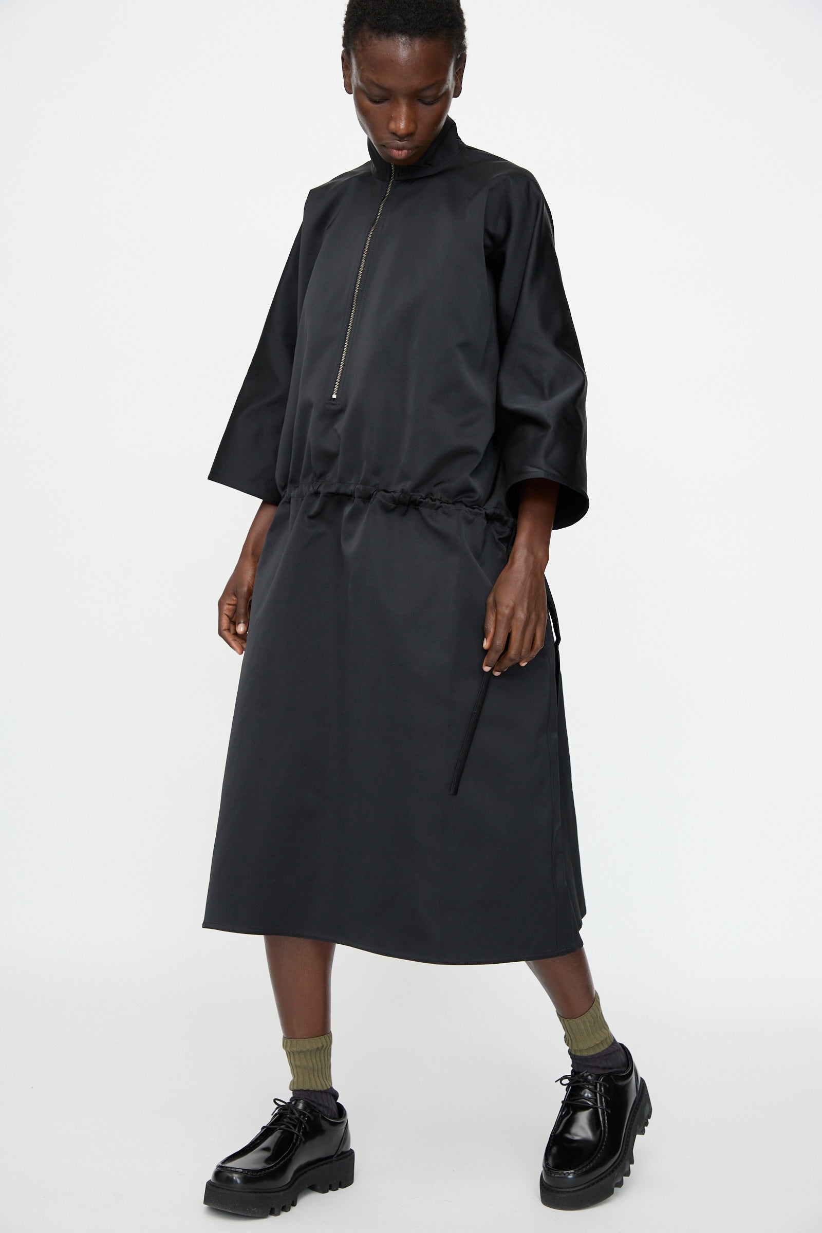 A person wears the Sofie D'Hoore Satin Dolci Dress in Black, styled with olive green socks and black platform shoes, standing against a plain white background.