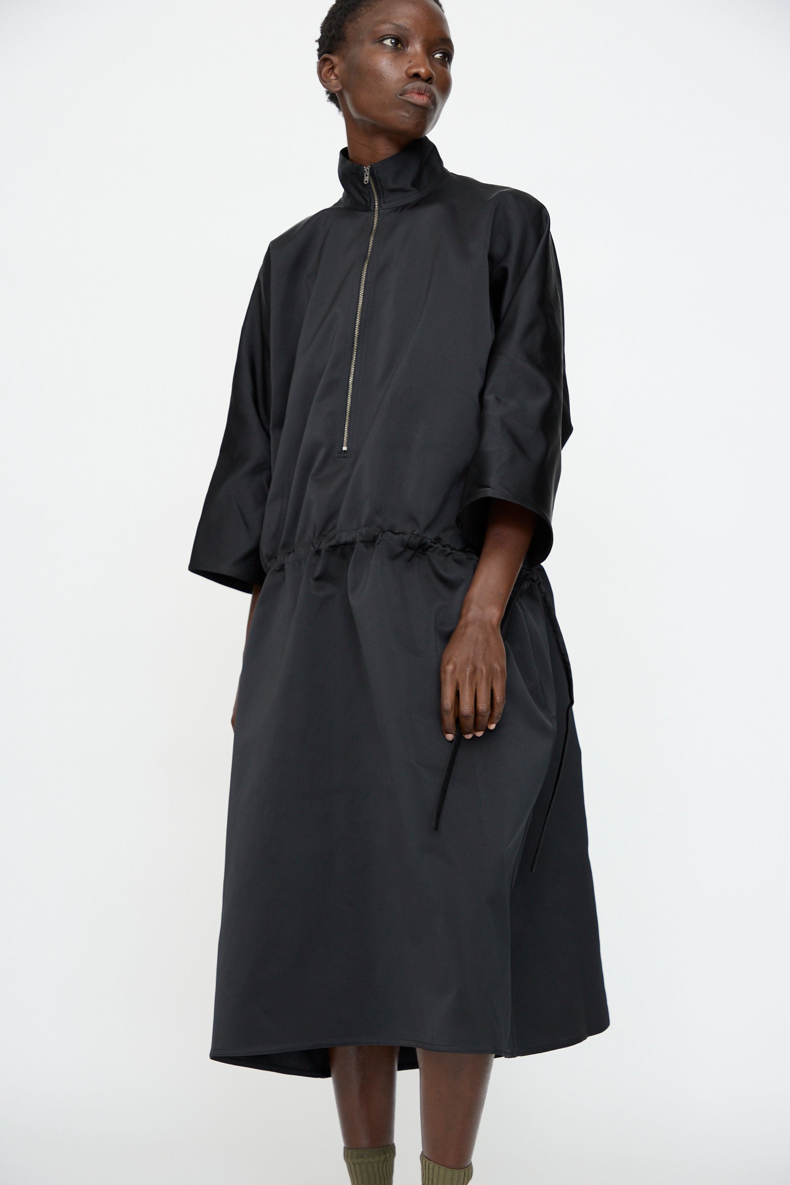 A person wearing the Sofie D'Hoore Satin Dolci Dress in Black, a loose-fitting midi dress with a zip-up collar and drawstring waist in satin cuir polyester, stands against a plain white background.