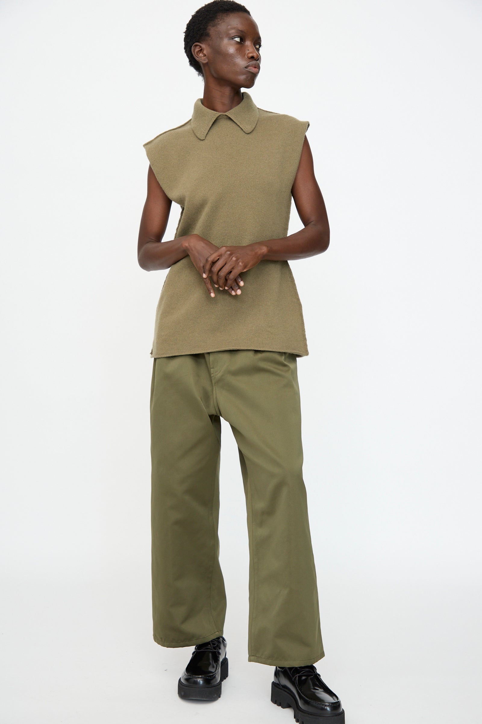 A person stands against a plain backdrop wearing a sleeveless tan top with a pointed collar, Sofie D'Hoore's Satin Polke Pant in Laurel, and black platform shoes.