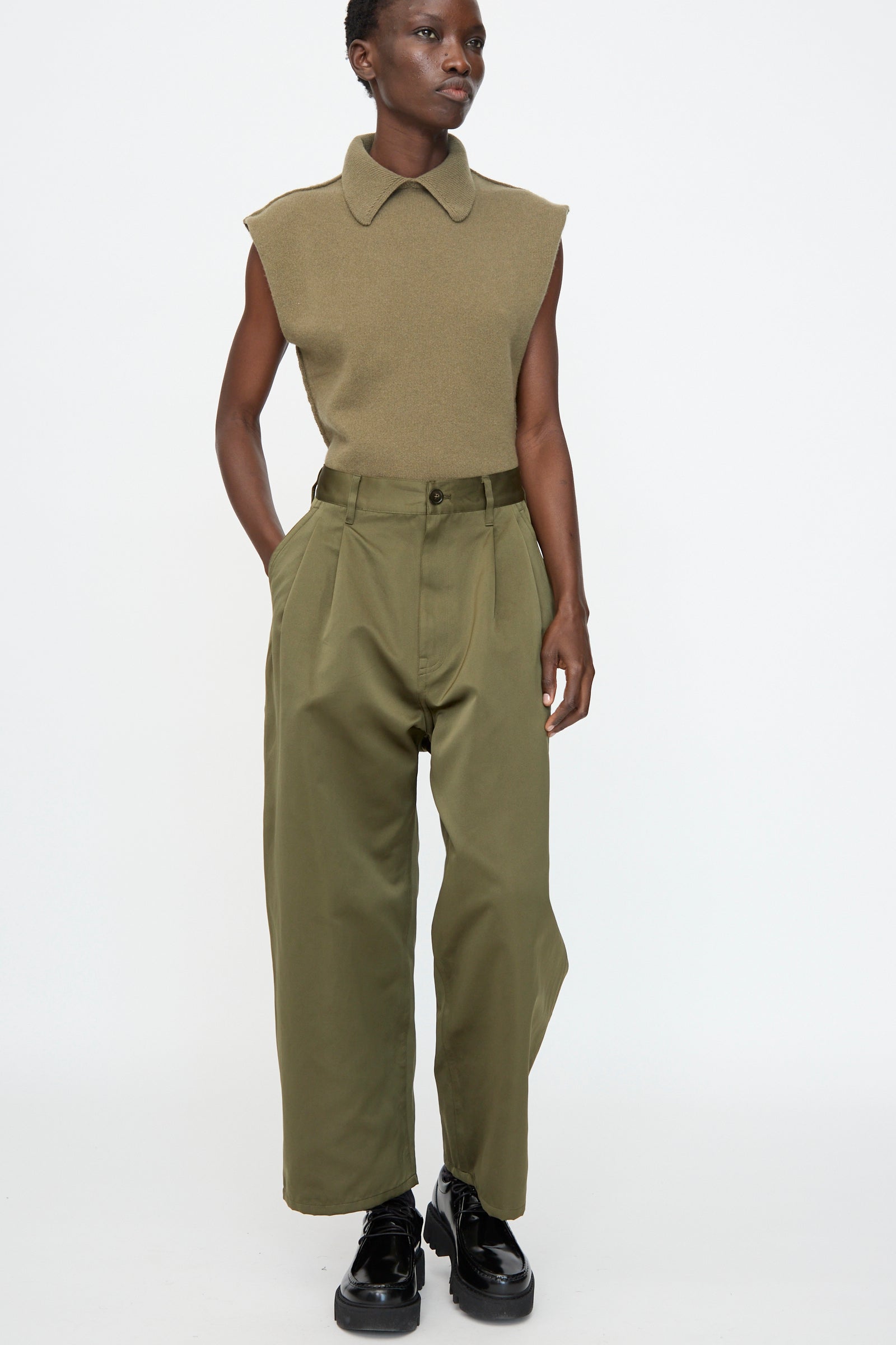 Person wearing a sleeveless olive-green knit top, Sofie D'Hoore Satin Polke Pant in Laurel, and black shoes, standing against a plain white background.
