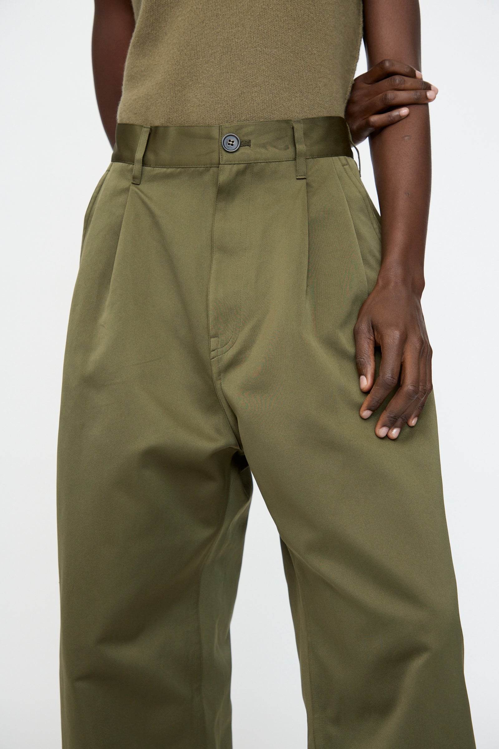 A person wears the Sofie D'Hoore Satin Polke Pant in Laurel, a high-waisted, wide-leg olive green trouser with an oversized work pant style, paired with an olive sleeveless top and one arm bent across their body.