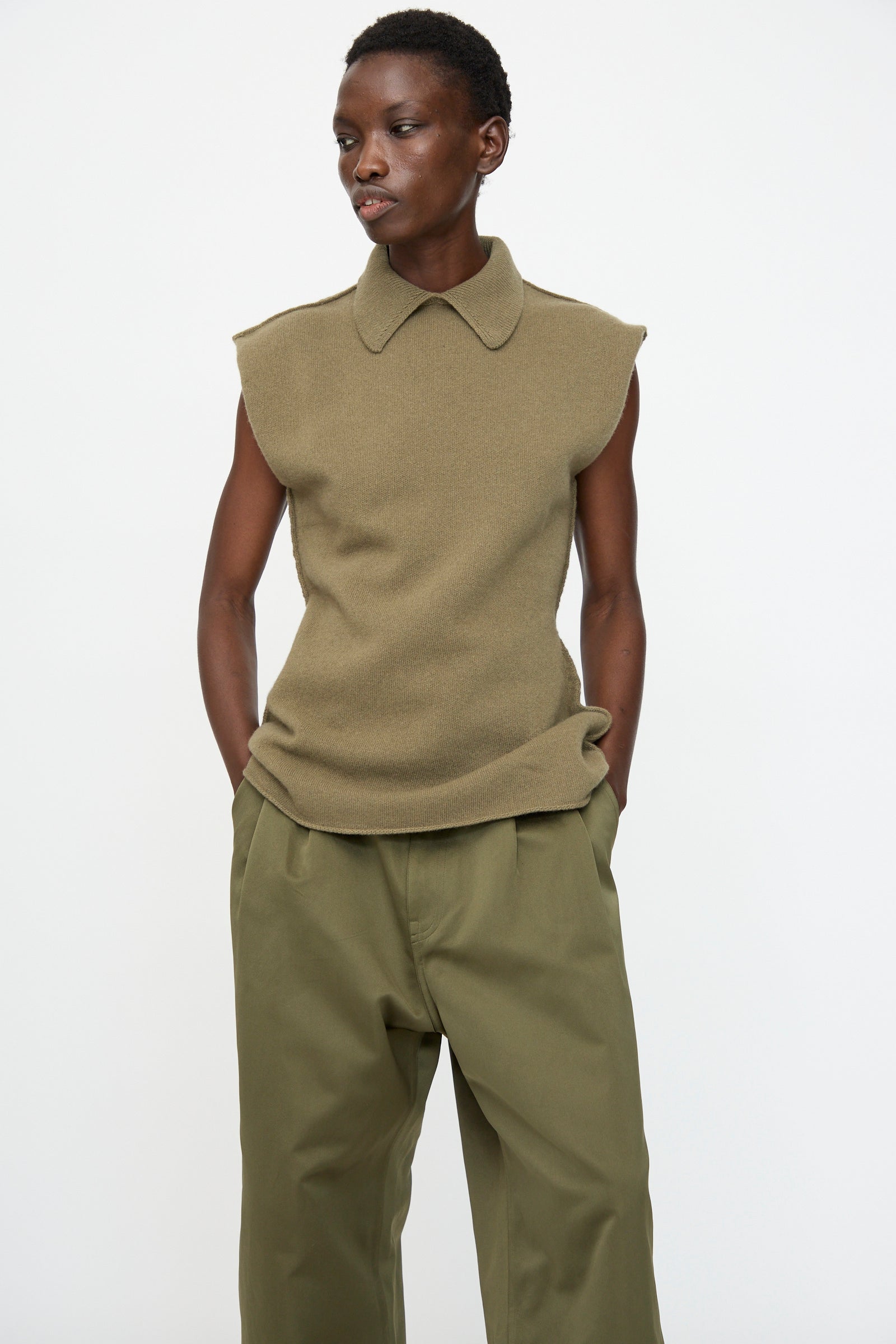 A person wears the Sofie D'Hoore Superfine Wool May Sleeveless Sweater in Sage with loose-fitting olive trousers, standing against a plain white background.