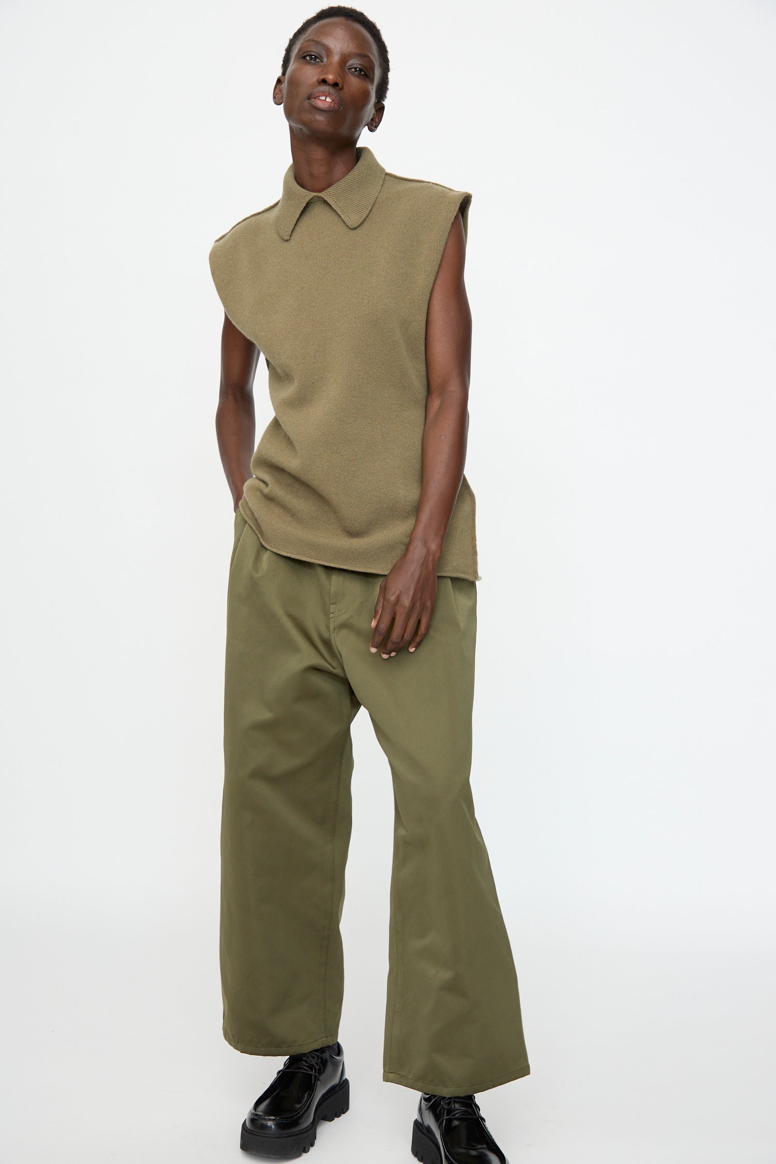 A person wears the Sofie D'Hoore Superfine Wool May Sleeveless Sweater in Sage, olive wide-leg pants, and black platform shoes, standing against a plain white background.