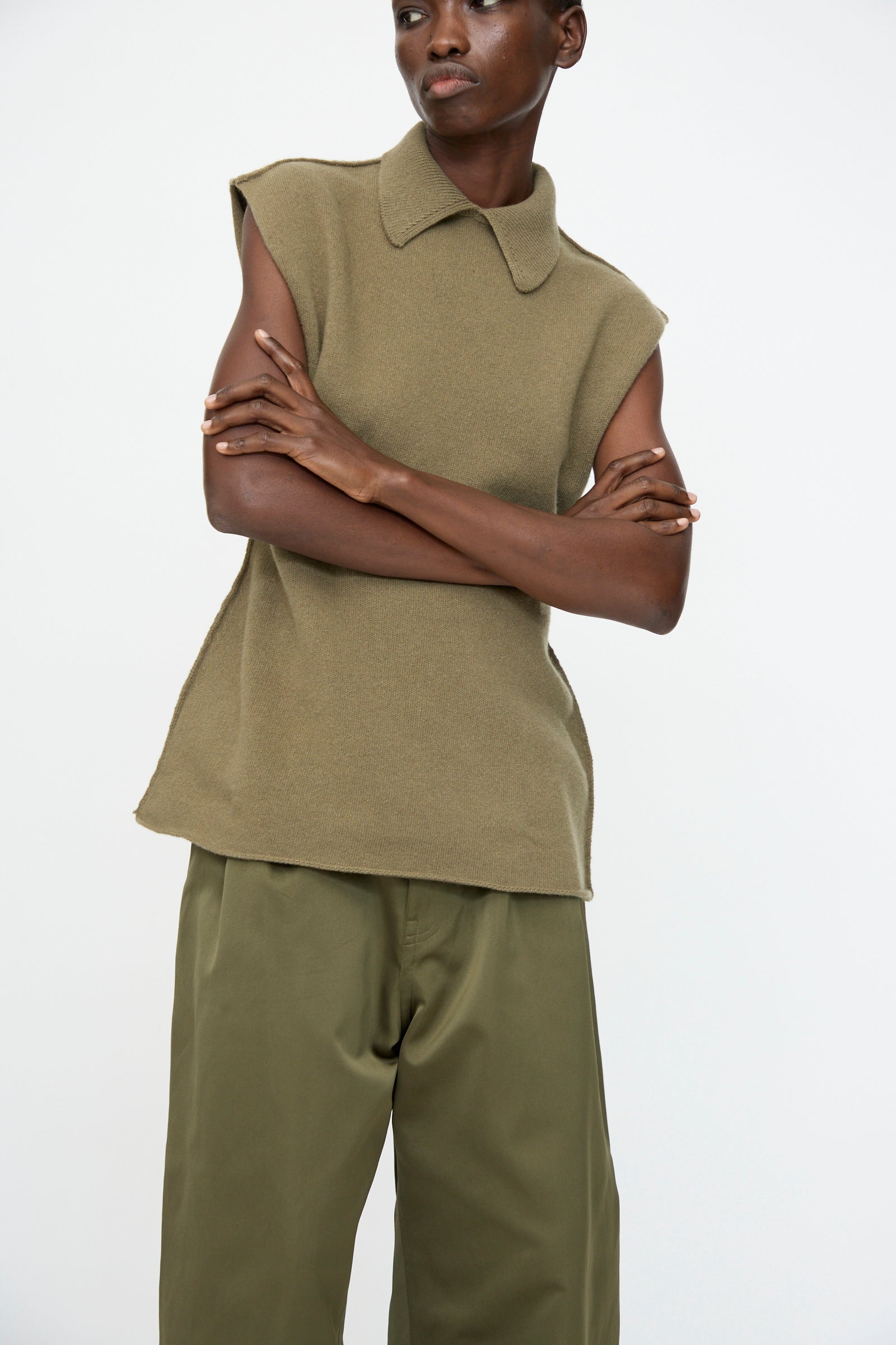 Wearing the Sofie D'Hoore Superfine Wool May Sleeveless Sweater in Sage, styled with loose olive green pants for an hourglass silhouette, arms crossed against a plain white background.