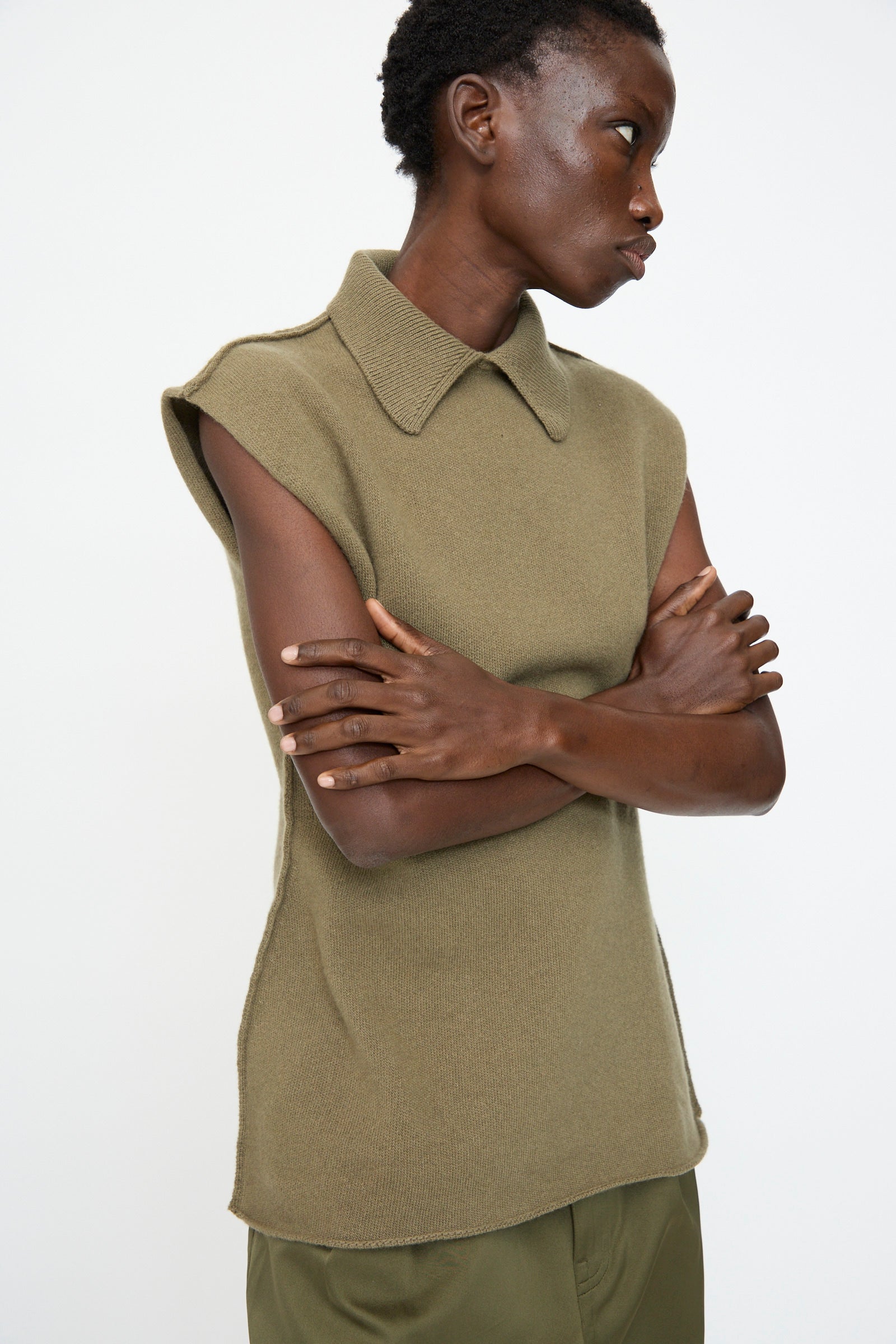 A person stands with arms crossed, wearing the Sofie D'Hoore Superfine Wool May Sleeveless Sweater in Sage, paired with matching trousers, highlighting an hourglass silhouette against a plain white background.