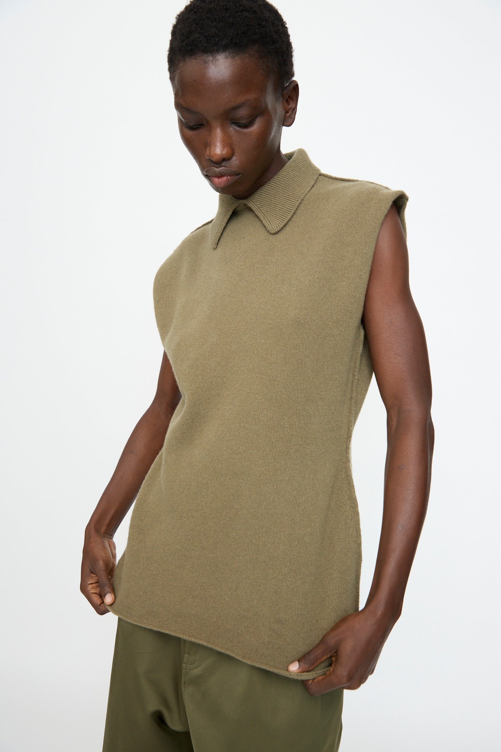 A person wears the Sofie D'Hoore Superfine Wool May Sleeveless Sweater in Sage with olive pants, standing against a plain white background and looking down.