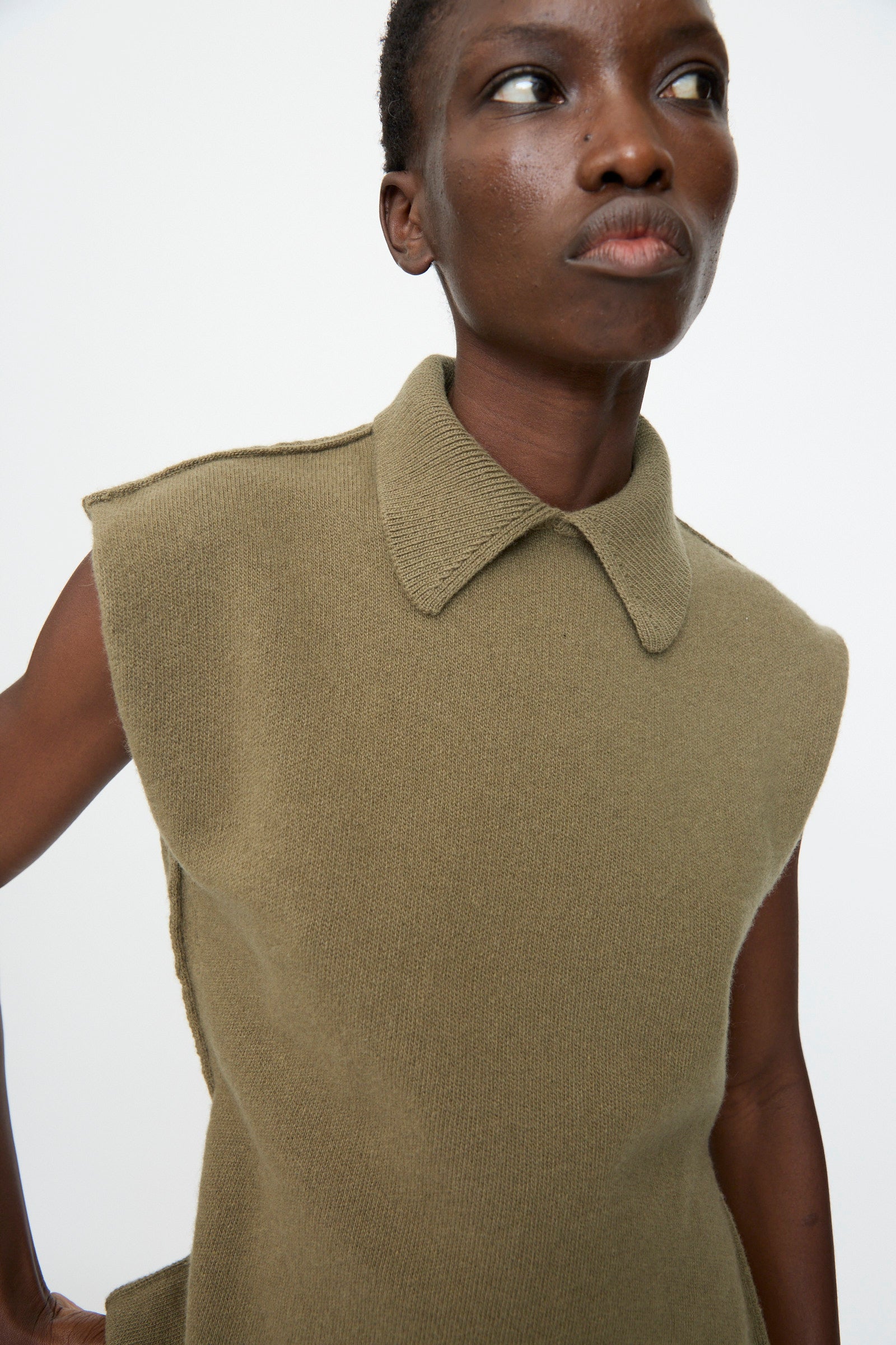 A person wears the Sofie D'Hoore Superfine Wool May Sleeveless Sweater in Sage, featuring a collar, while standing against a plain white background and looking slightly to the side.