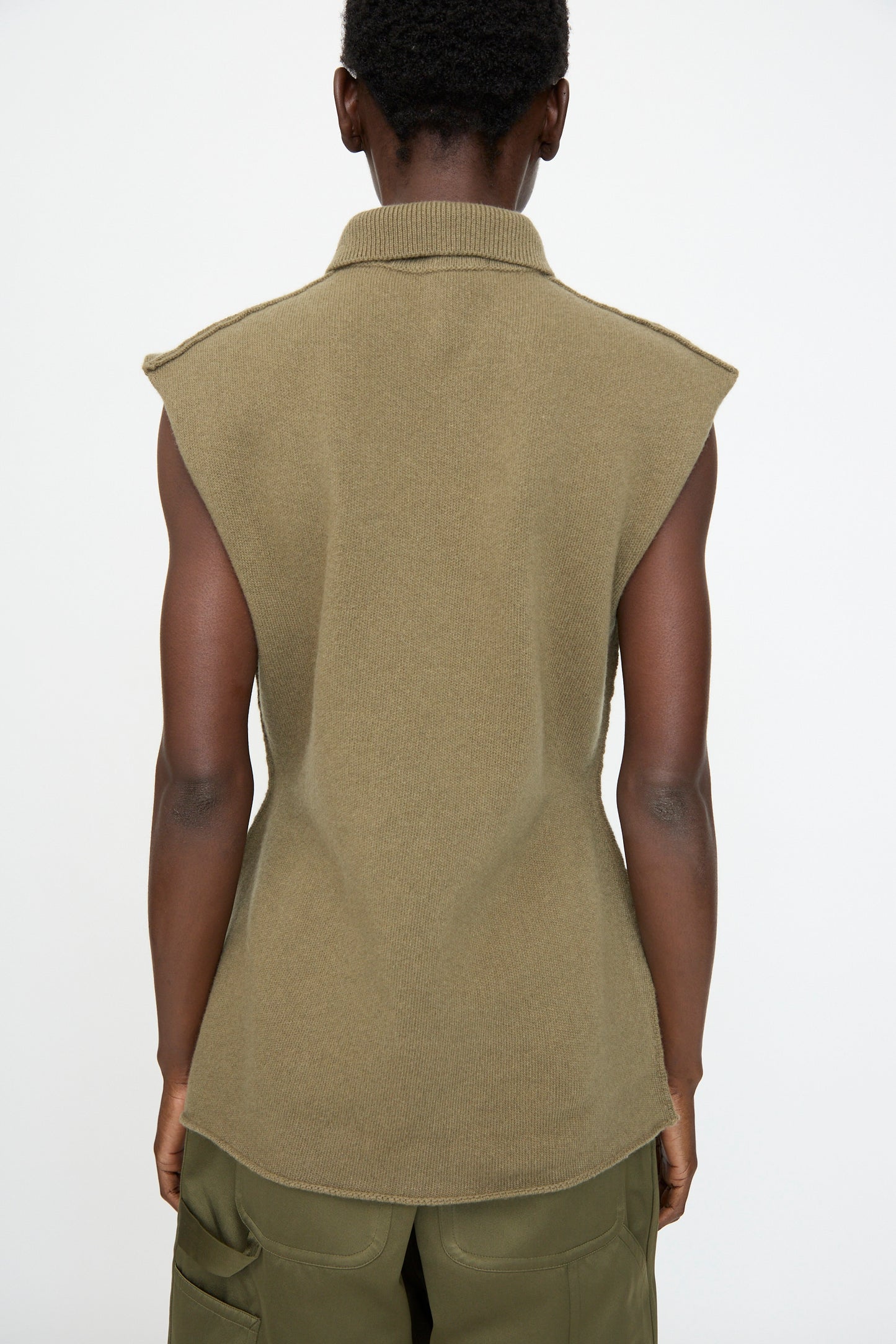 A person stands with their back to the camera, wearing the Sofie D'Hoore Superfine Wool May Sleeveless Sweater in Sage and green cargo pants, highlighting an hourglass silhouette against a plain background.
