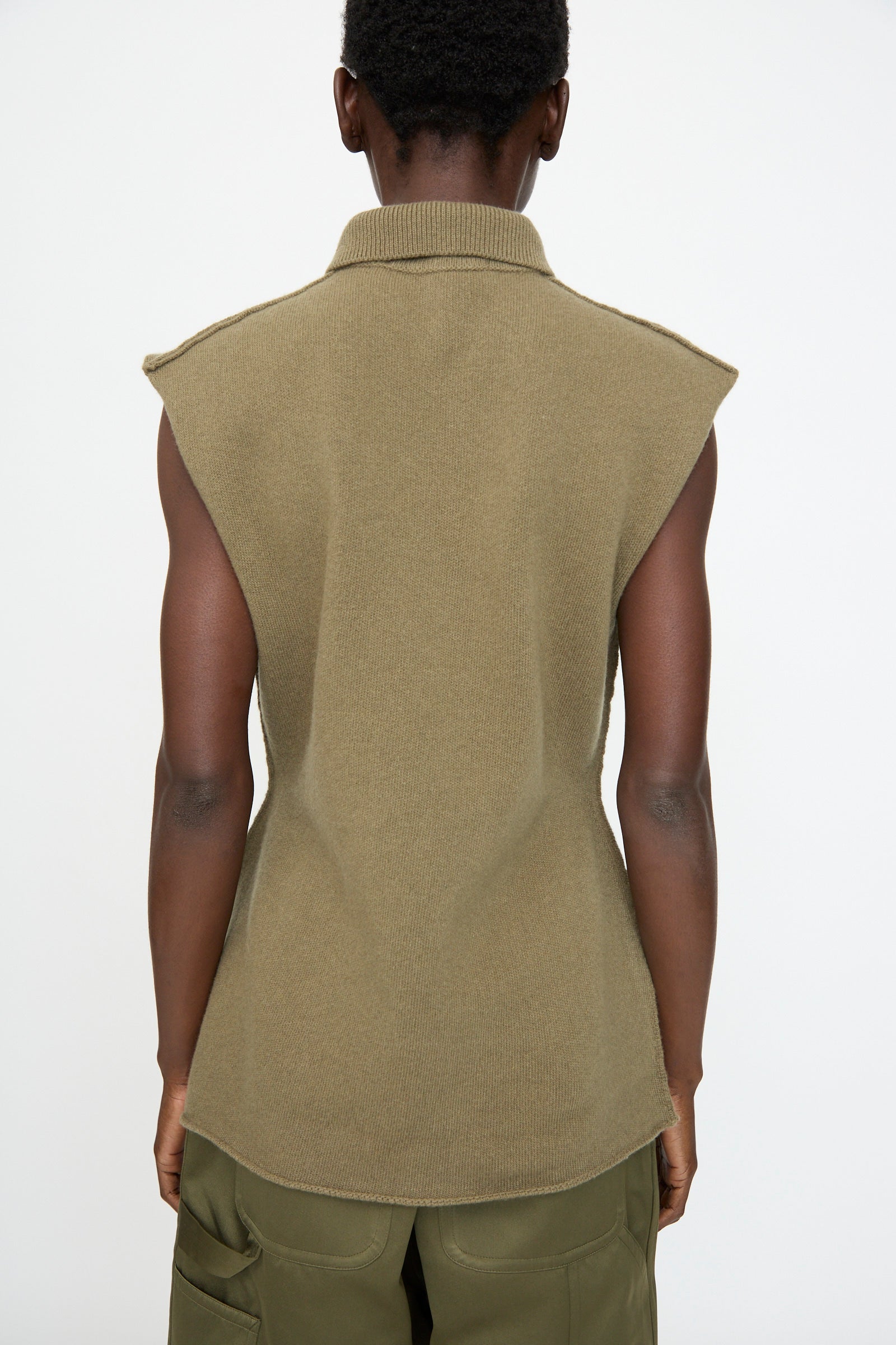A person stands with their back to the camera, wearing the Sofie D'Hoore Superfine Wool May Sleeveless Sweater in Sage and green cargo pants, highlighting an hourglass silhouette against a plain background.