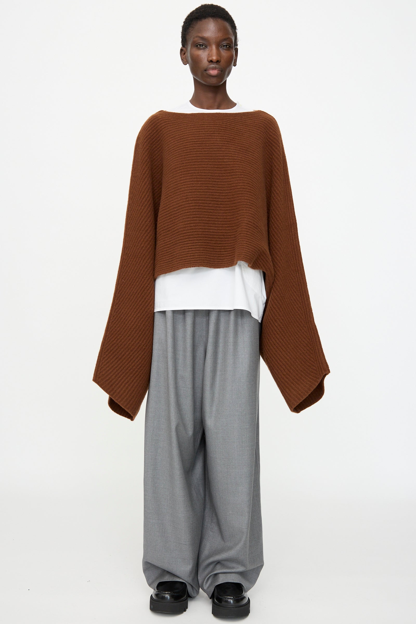 A person wears the Sofie D'Hoore Superfine Wool Murakami Oversized Sweater in Chestnut over a white shirt, paired with gray wide-leg trousers and black shoes, standing against a plain white background.