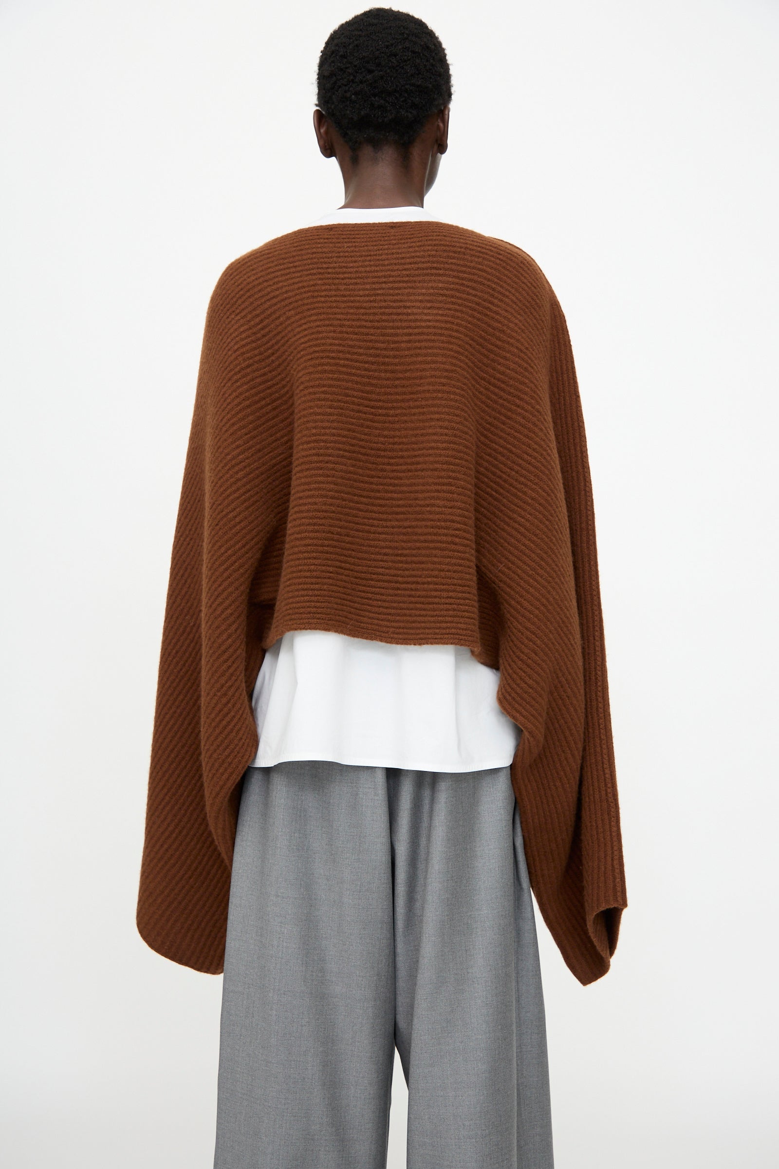 A person stands with their back to the camera, wearing the Sofie D'Hoore Superfine Wool Murakami Oversized Sweater in Chestnut over a white top and gray wide-leg pants, set against a plain background.