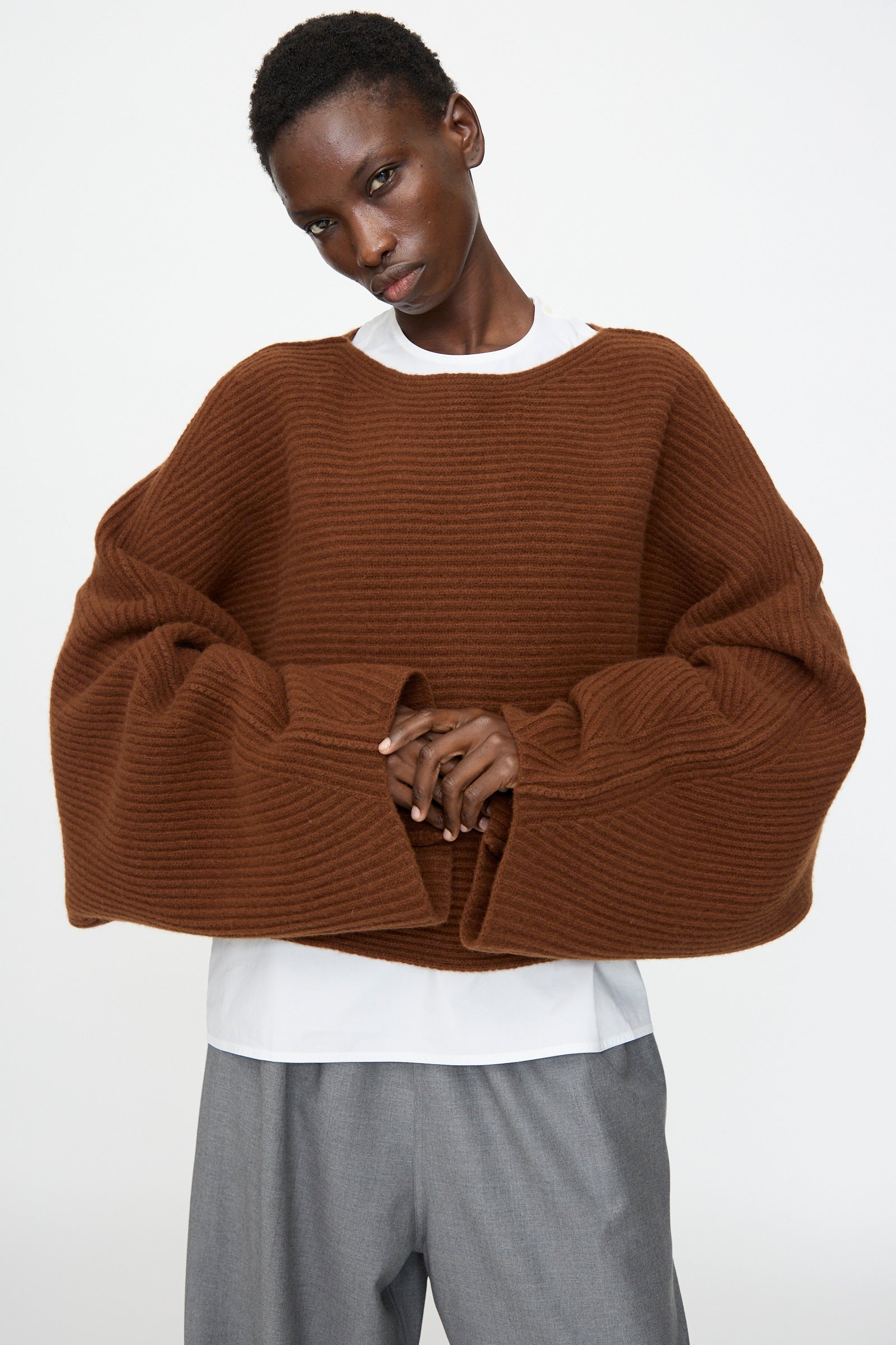 A person wears the Sofie D'Hoore Superfine Wool Murakami Oversized Sweater in Chestnut over a white shirt and gray pants, standing against a plain background.