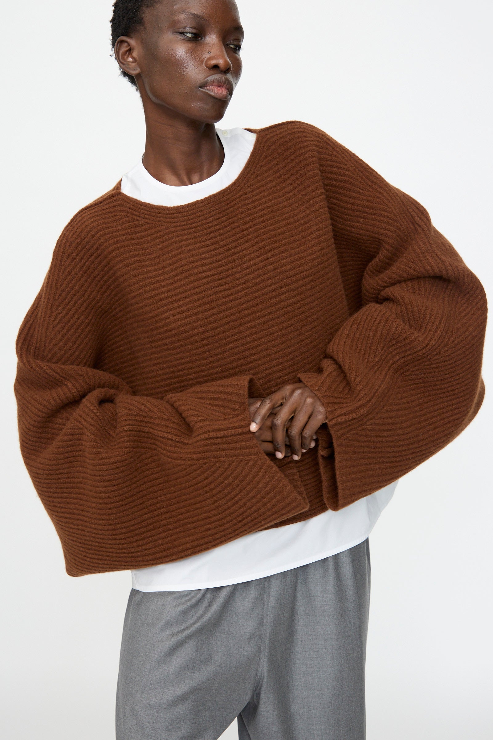 Person wearing the Sofie D'Hoore Superfine Wool Murakami Oversized Sweater in Chestnut over a white shirt with gray trousers, standing against a plain background.