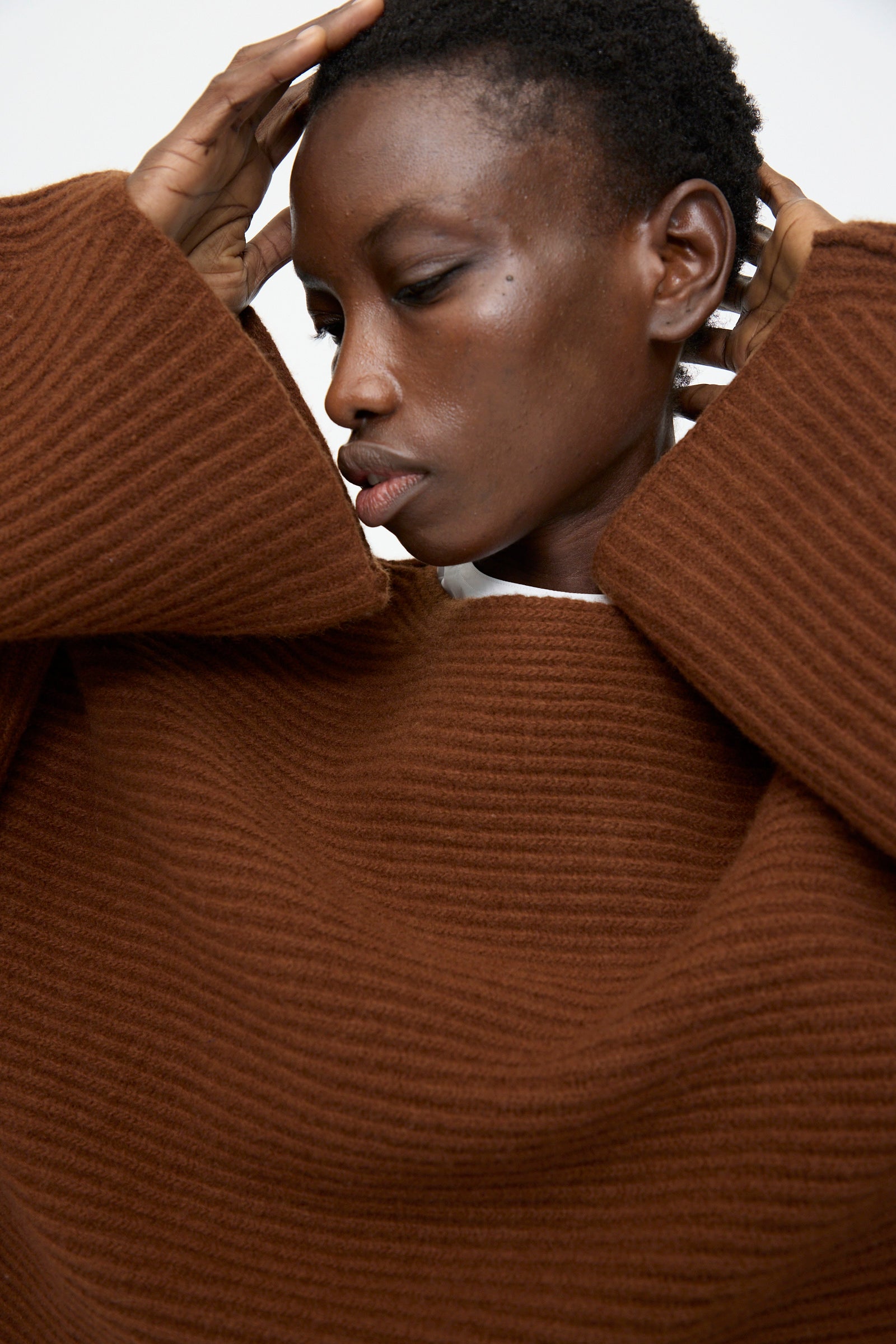 A model in the Sofie D'Hoore Superfine Wool Murakami Oversized Sweater in Chestnut stands with hands on head, gazing downward against a plain background.