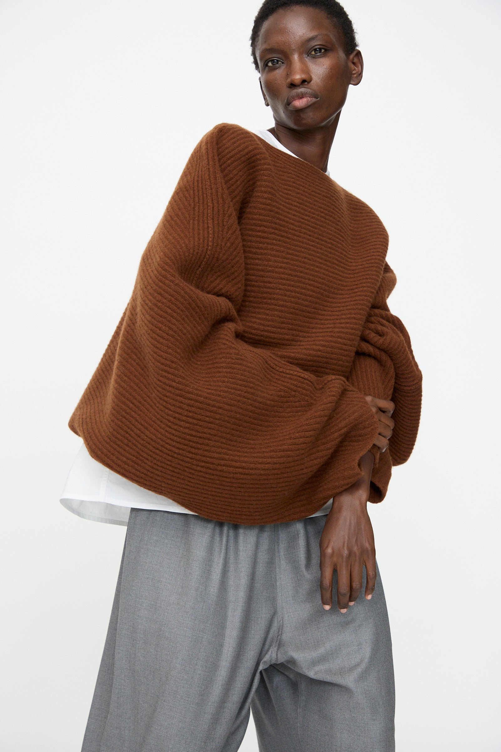 Model wears the Sofie D'Hoore Superfine Wool Murakami Oversized Sweater in Chestnut with wide sleeves, styled with loose gray trousers against a plain light backdrop.