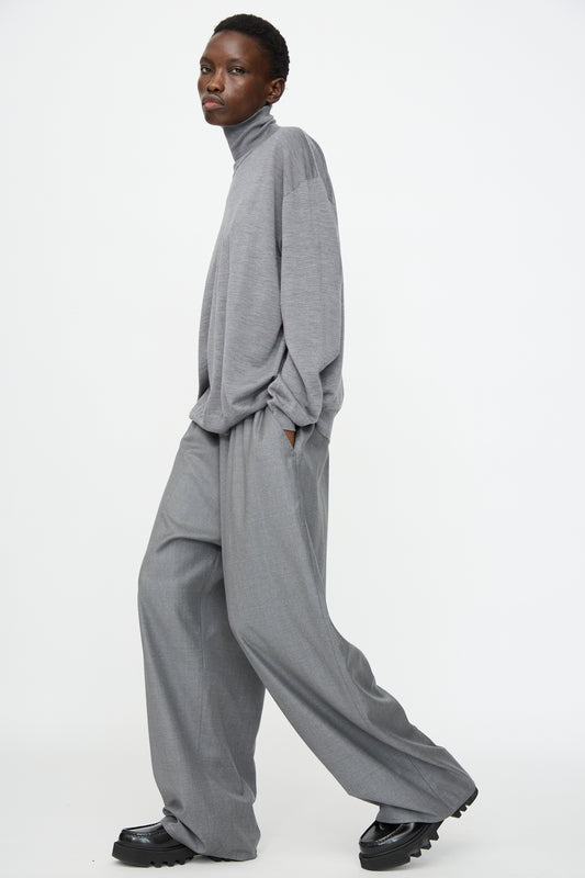 A person wearing Sofie D'Hoore's Superfine Wool Pasch Pants in Mid Grey Melange, paired with a loose gray turtleneck and black shoes, stands against a plain white background.