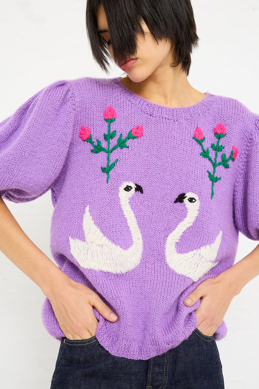 Woman in a Sofio Gongli hand-knit Swans Sweater in Purple featuring white swan embroidery and red floral details, with her hands on her hips.