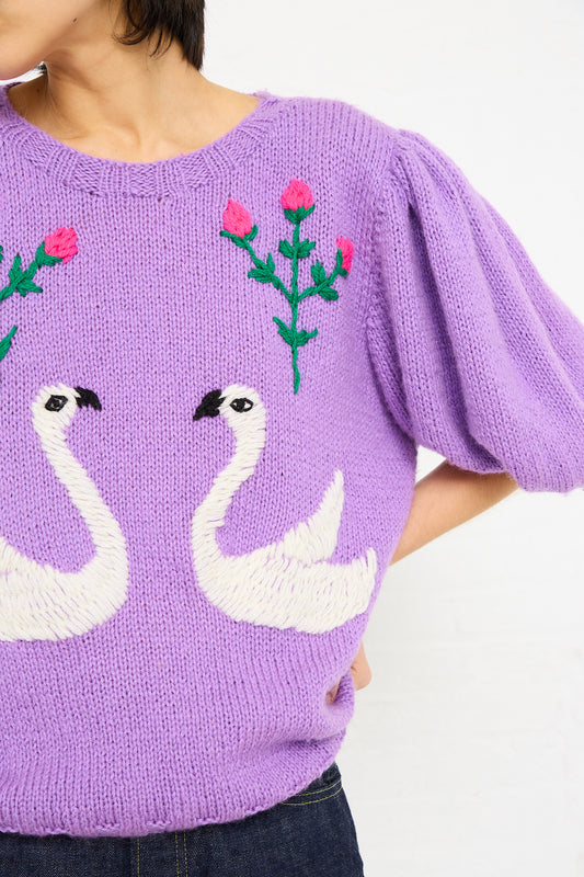 A person in a Sofio Gongli lavender Swans Sweater featuring white swan embroidery on the front, with pink floral details. Only the torso and arms are visible.