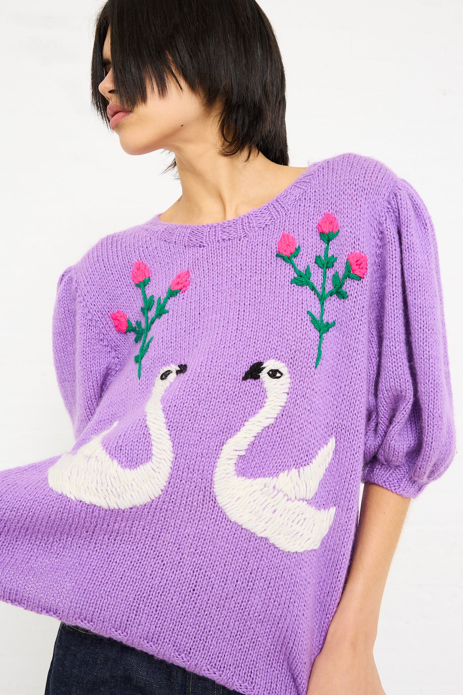 Woman in a Sofio Gongli hand-knit sweater with embroidered swans and flowers, profile view, against a white background.