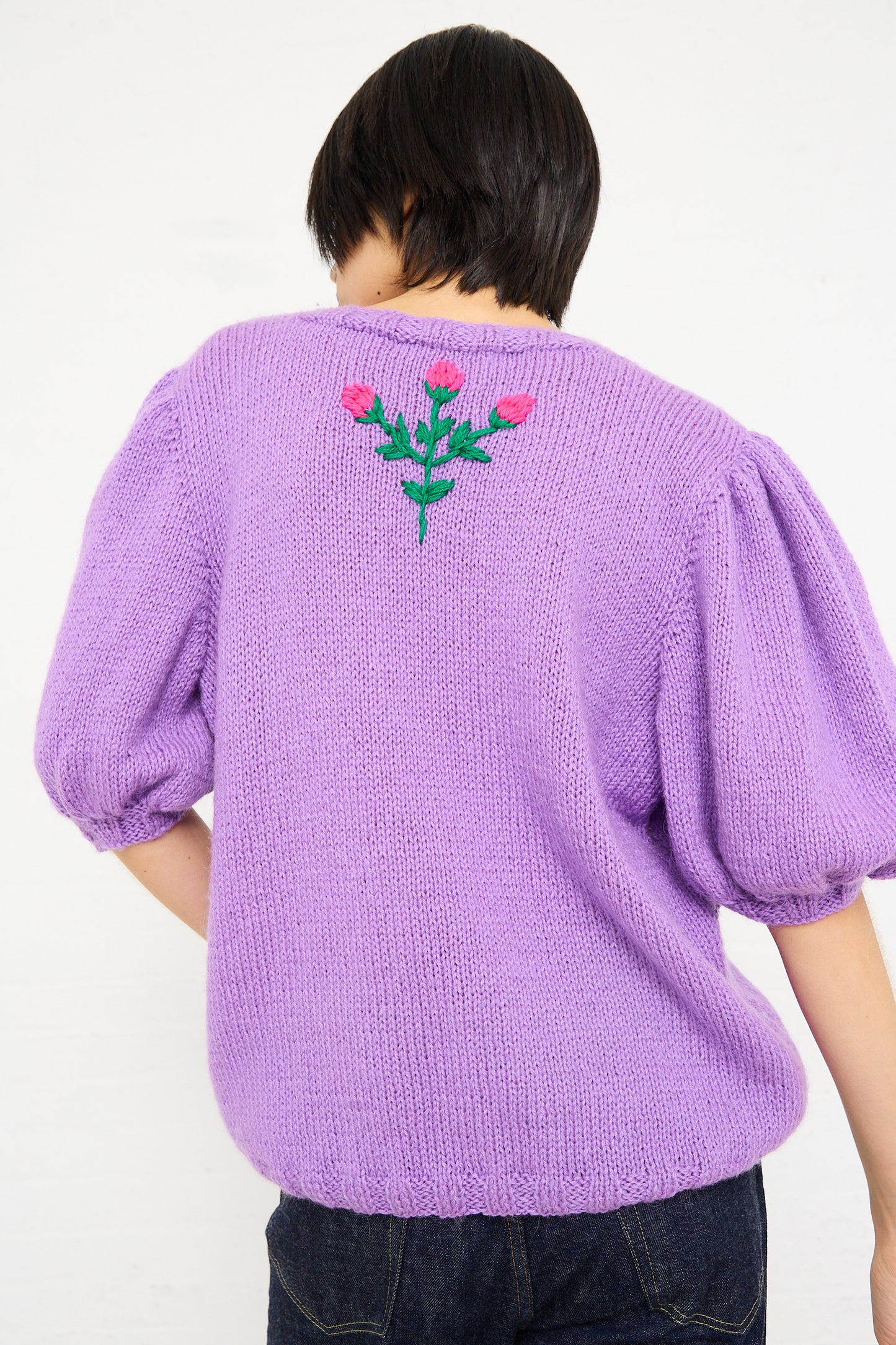Rear view of a woman wearing a Sofio Gongli Swans Sweater in Purple with floral embroidery on the back, standing against a white background.
