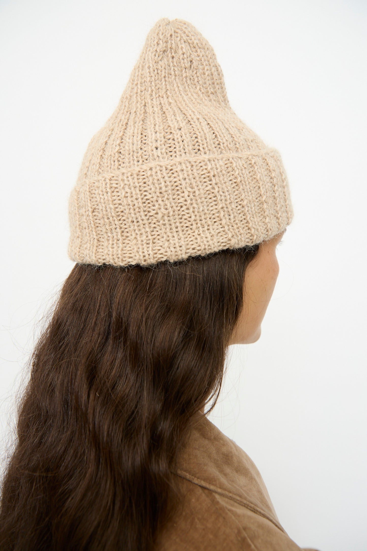 Person with long wavy brown hair wearing the SONO Alpaca and Wool Gyu Beanie in Oat and a brown jacket, shown from the back against a plain background.
