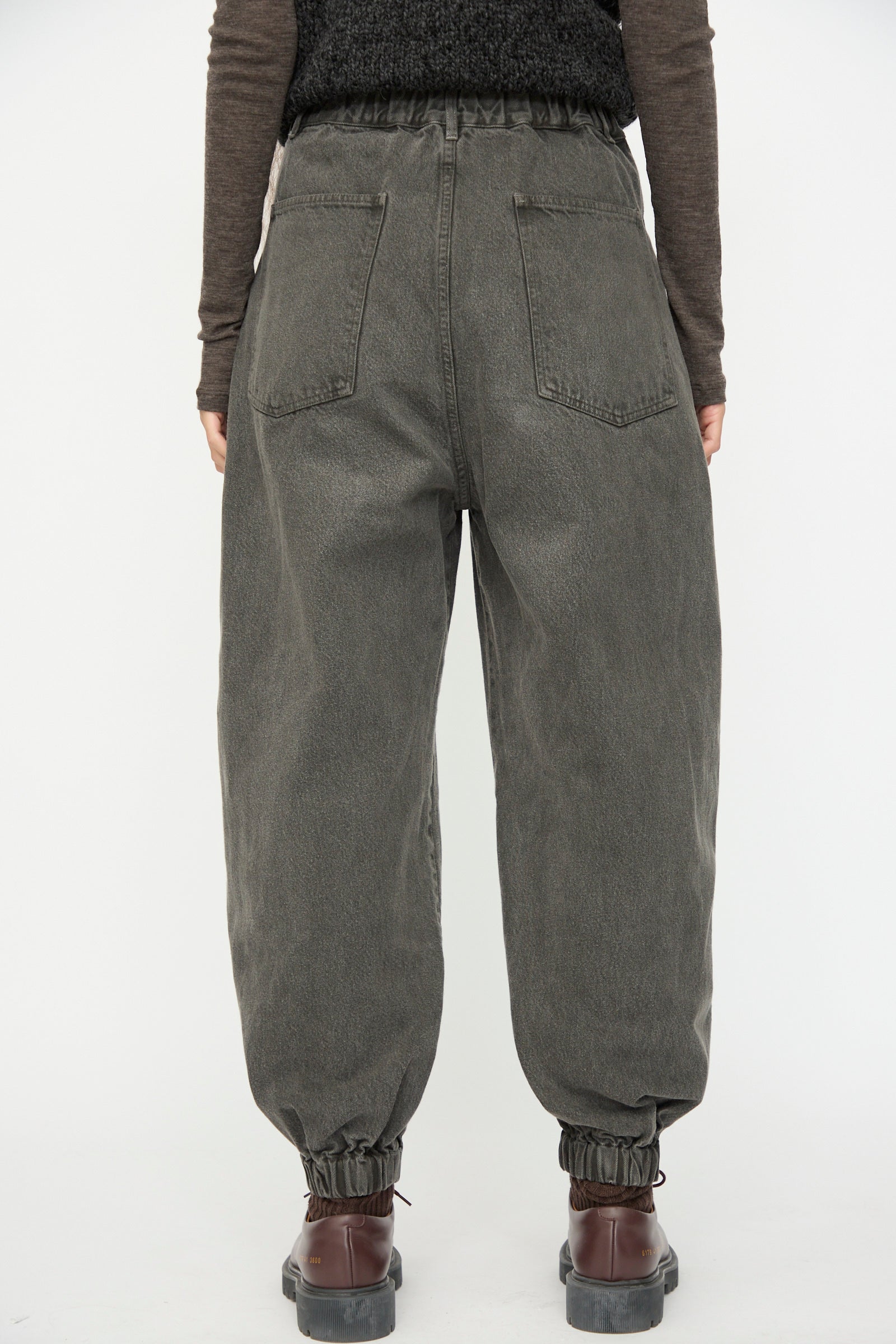 A person stands facing away against a plain white background, wearing SONO Cotton Jah Jeans in Off Black with an elasticated waist, paired with brown shoes and a long-sleeve brown top.
