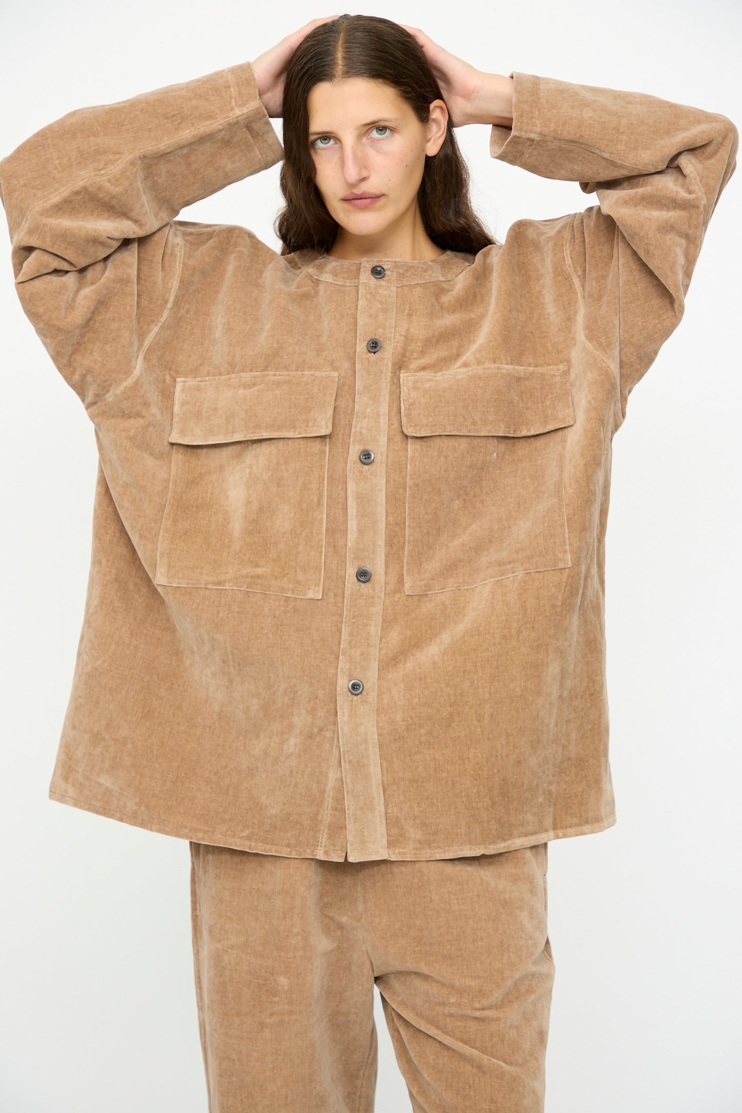 A person with long brown hair wears the SONO Sid Shirt in Desert Rose, a relaxed fit tan corduroy button-down with large front pockets and matching pants, standing against a plain white background.