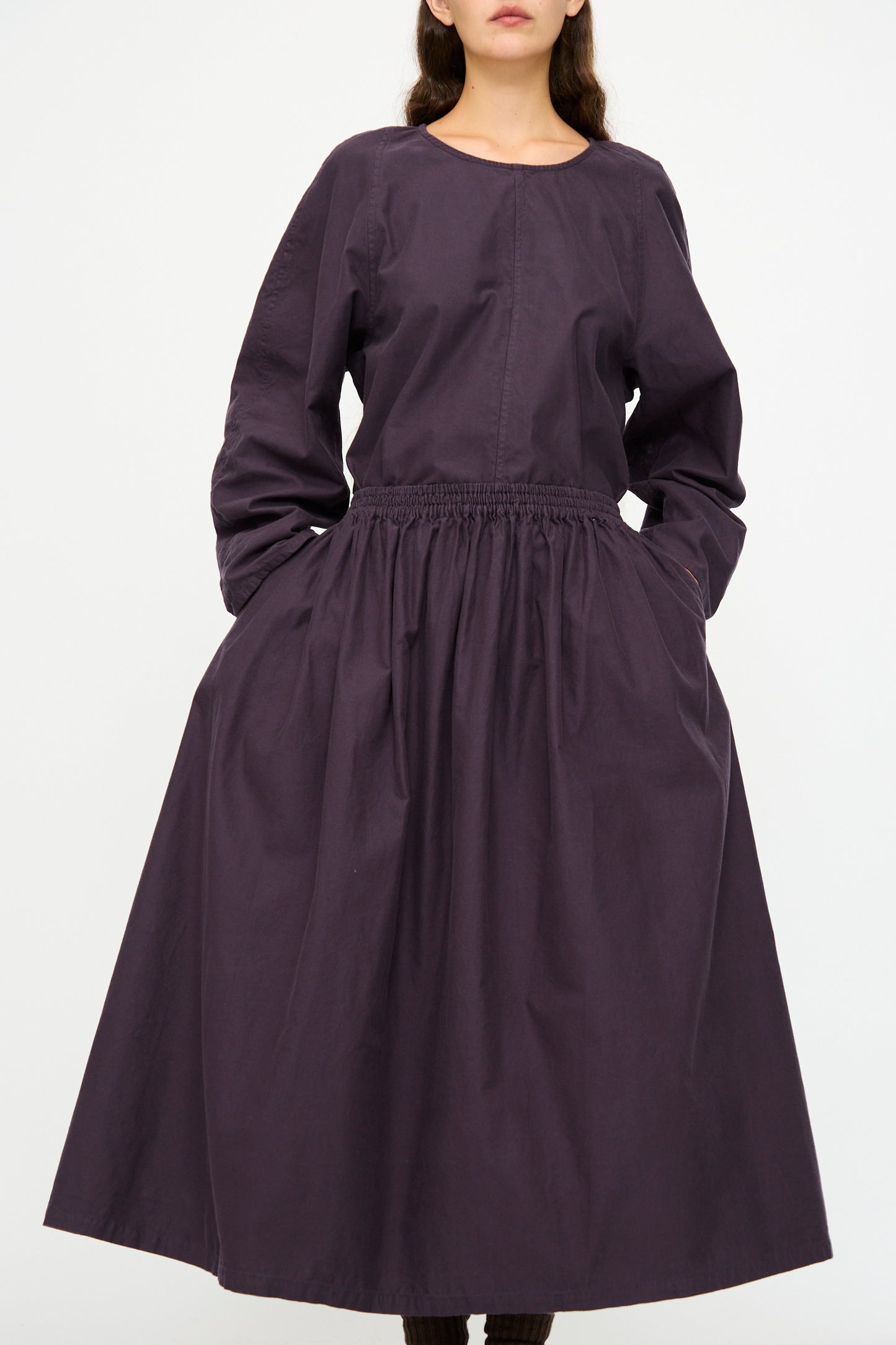 A woman models the Skye Skirt in Dark Grape by SONO, a long, pocketed skirt with a gathered waist and deep purple hue, standing against a plain white background.