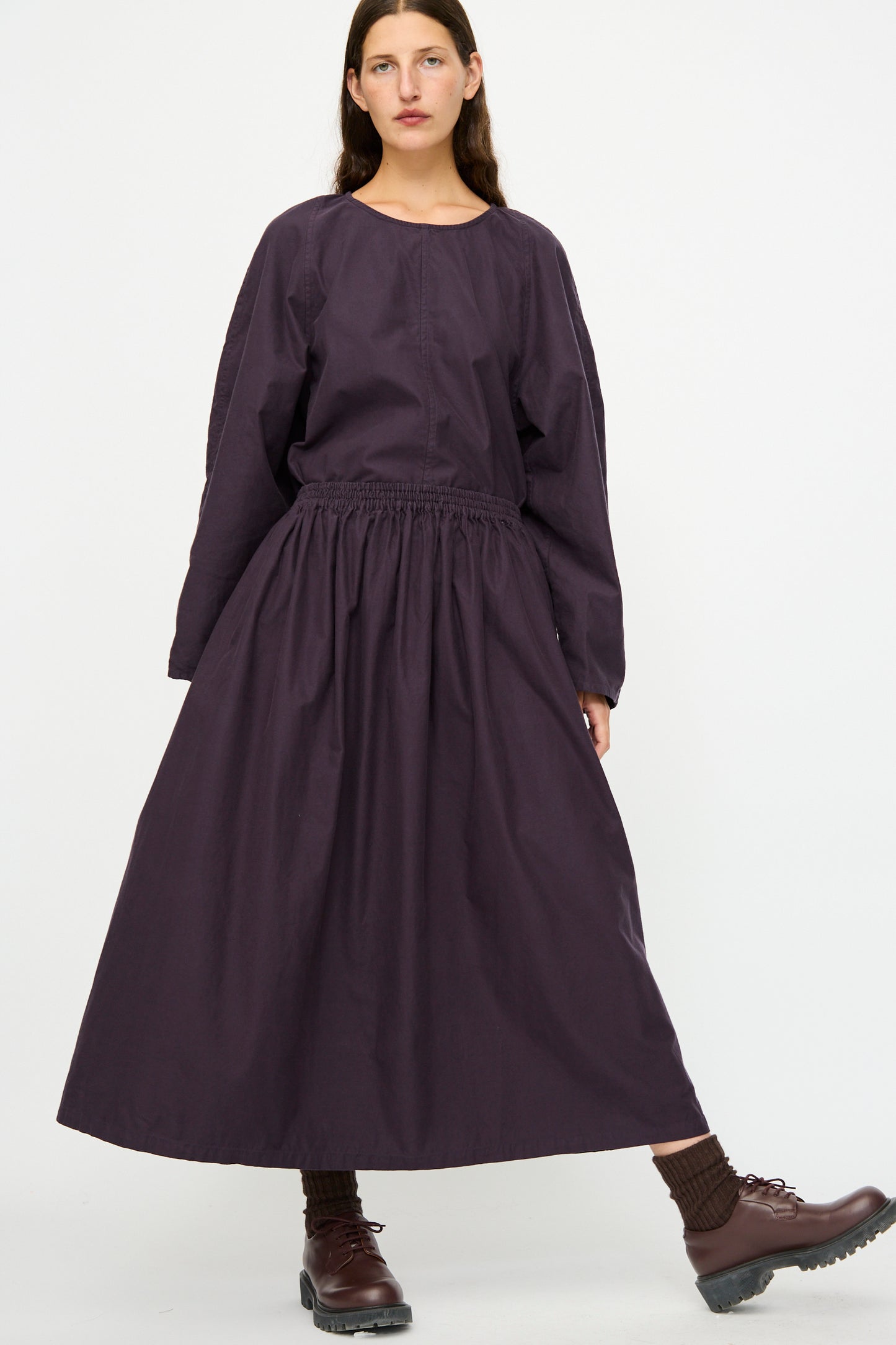 A person wears the SONO Skye Skirt in Dark Grape, styled with a loose dark purple dress, brown socks, and brown lace-up shoes, standing against a plain white background.