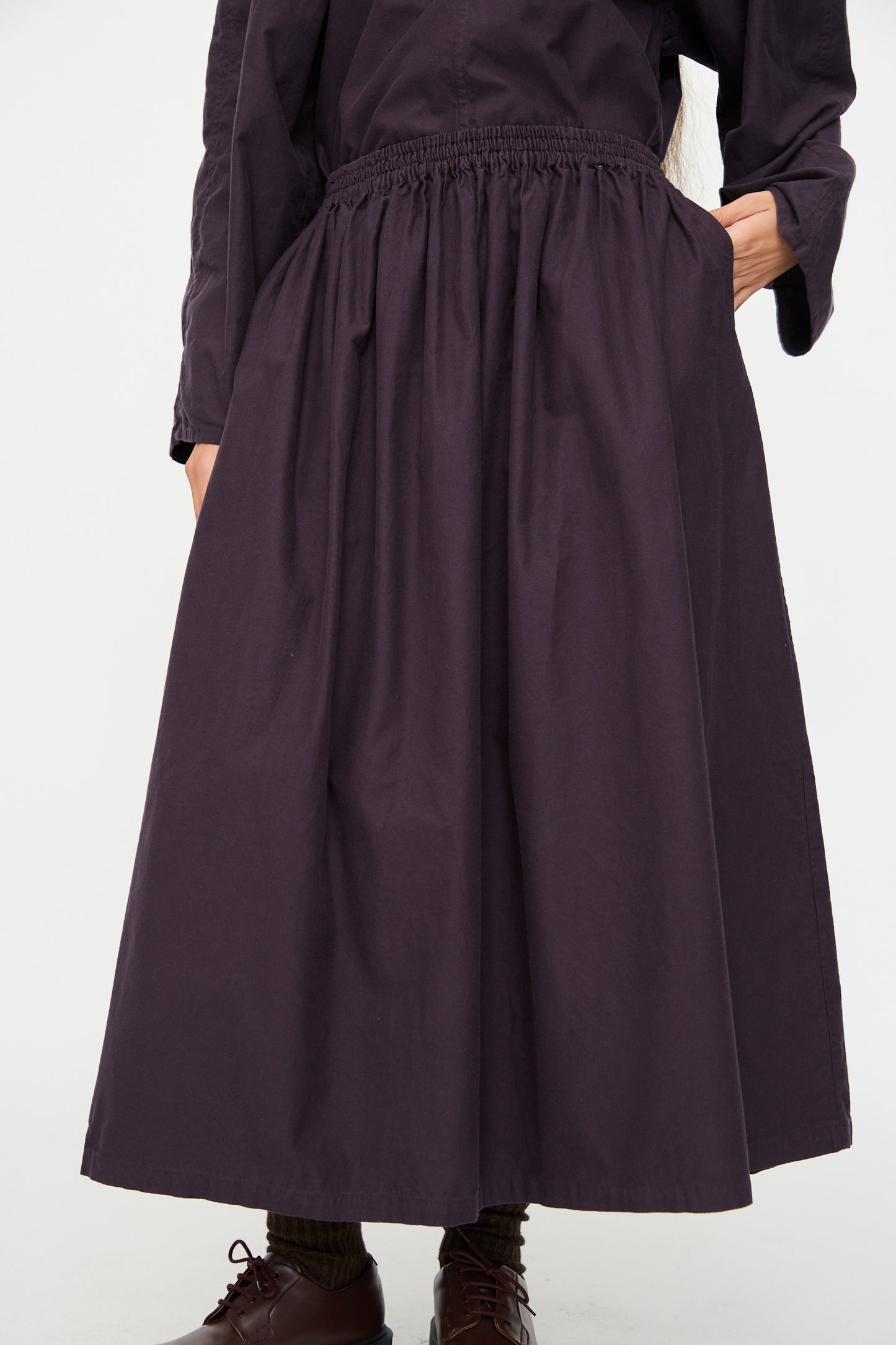 A person wearing the SONO Skye Skirt in Dark Grape, featuring organic cotton and an elastic waistband, paired with brown socks and brown shoes, stands with one hand in a pocket.