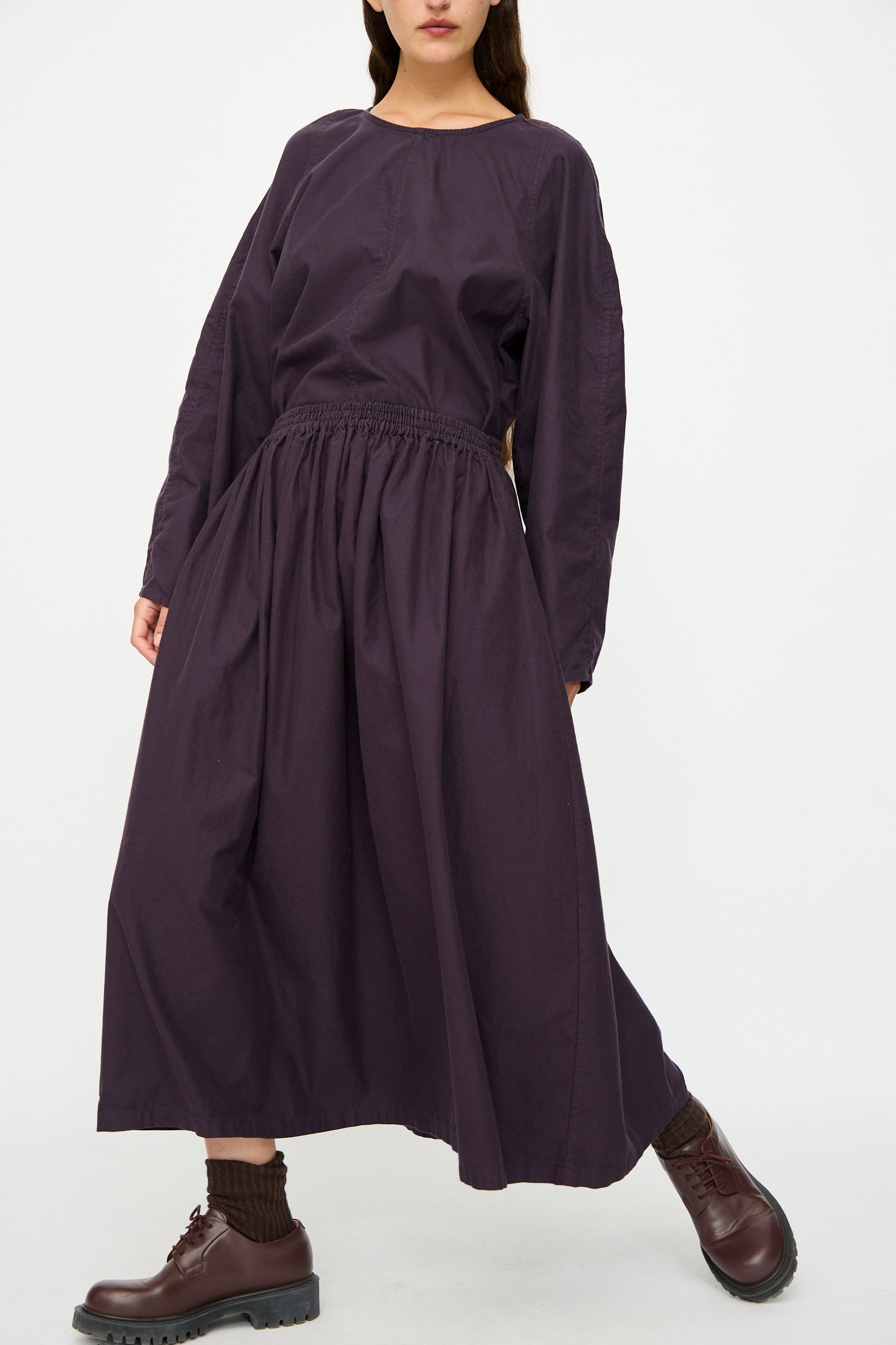 A woman wears the SONO Skye Skirt in Dark Grape, styled with brown socks and chunky brown shoes, standing against a plain white background.