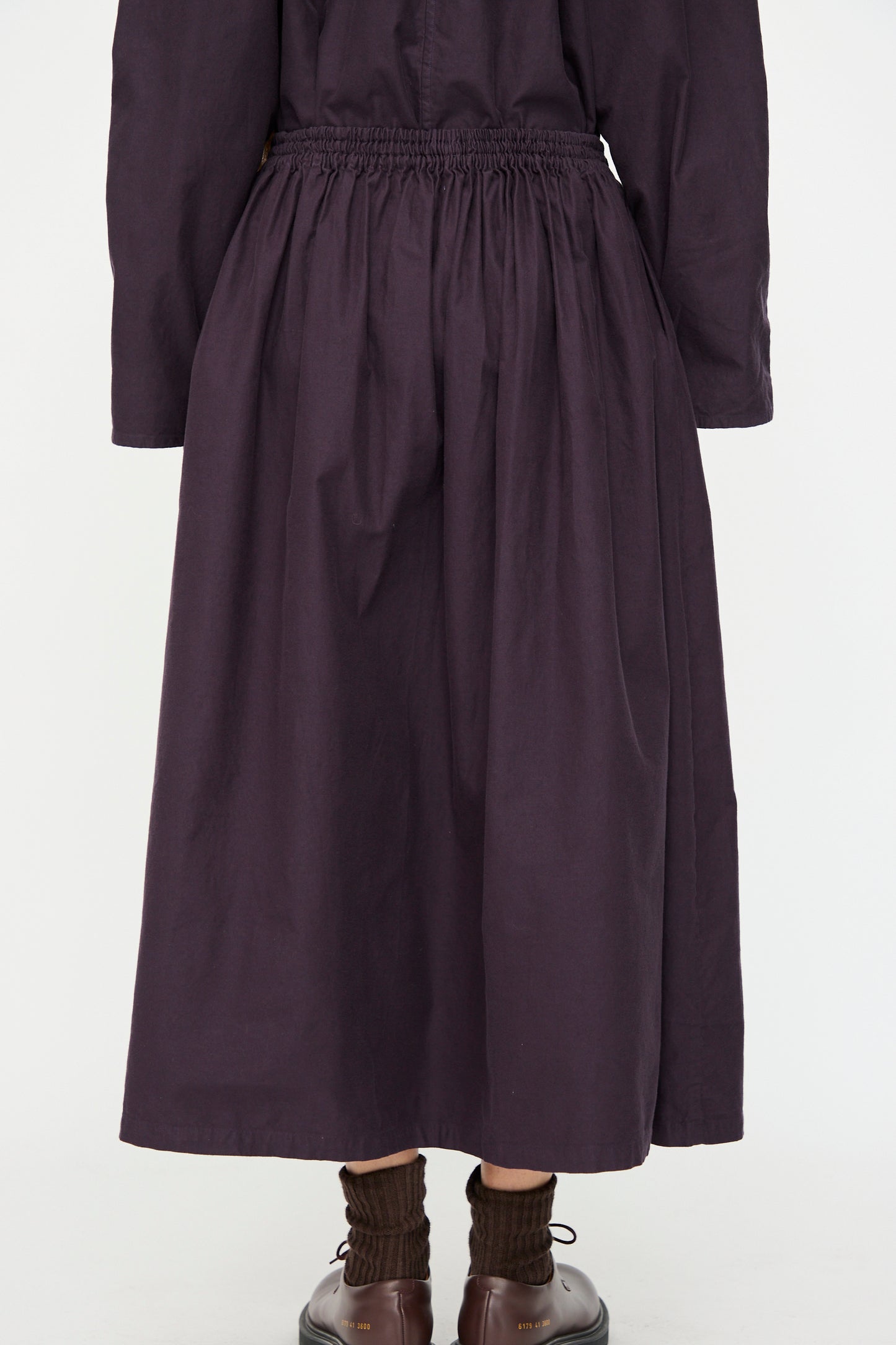 A person wears the SONO Skye Skirt in Dark Grape—an organic cotton skirt with an elastic waistband—paired with brown socks and shoes, shown from the back against a plain white background.