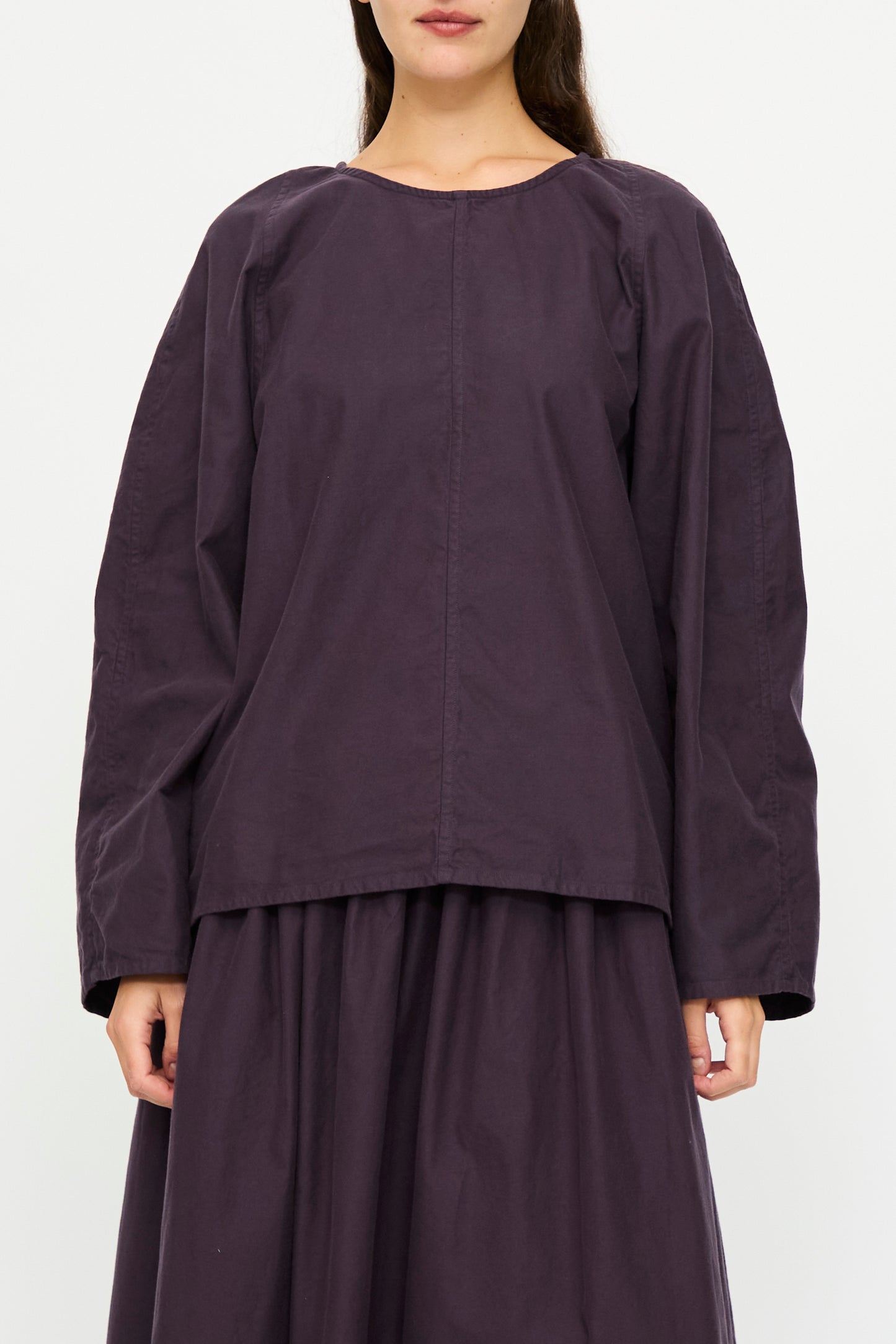 A woman stands against a plain white background wearing the SONO Tiga Top in Dark Grape, a loose-fitting, long-sleeved cotton poplin blouse paired with a matching skirt.