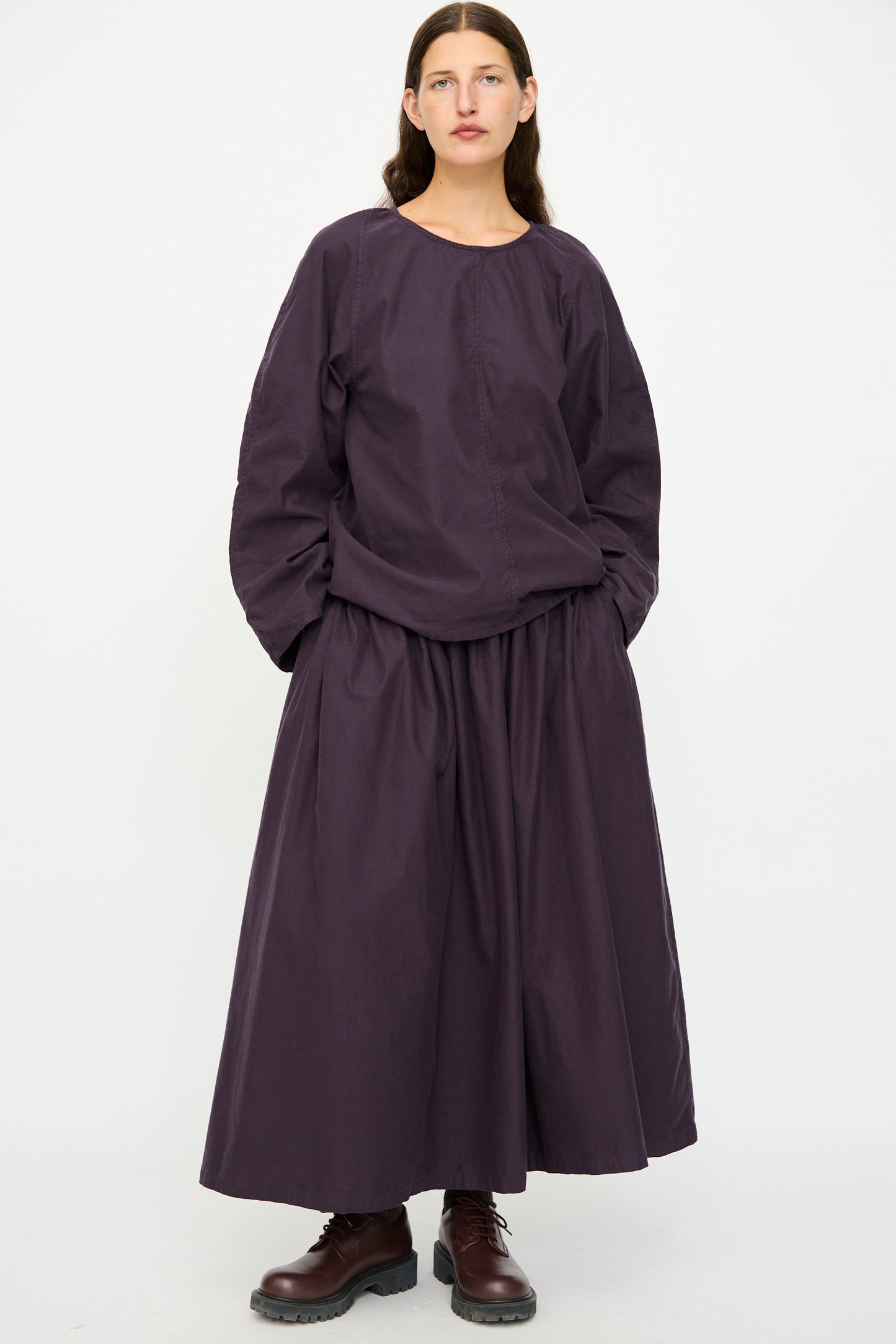 A person stands against a white background wearing the SONO Tiga Top in Dark Grape, paired with a matching full-length skirt and brown lace-up boots.
