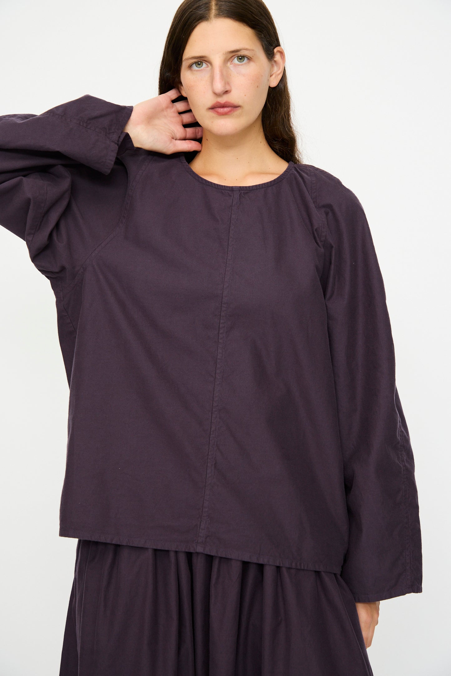 A woman with long brown hair wears the SONO Tiga Top in Dark Grape, styled with a skirt, standing against a plain white background.