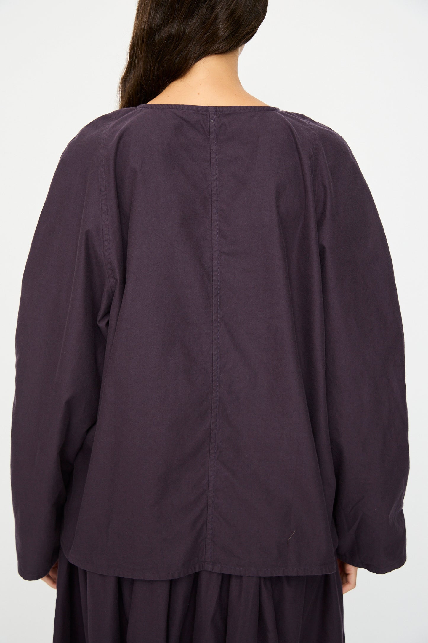 Person shown from behind wearing the SONO Tiga Top in Dark Grape, an oversized, long-sleeved organic cotton top with a center seam detail; long brown hair partially visible.