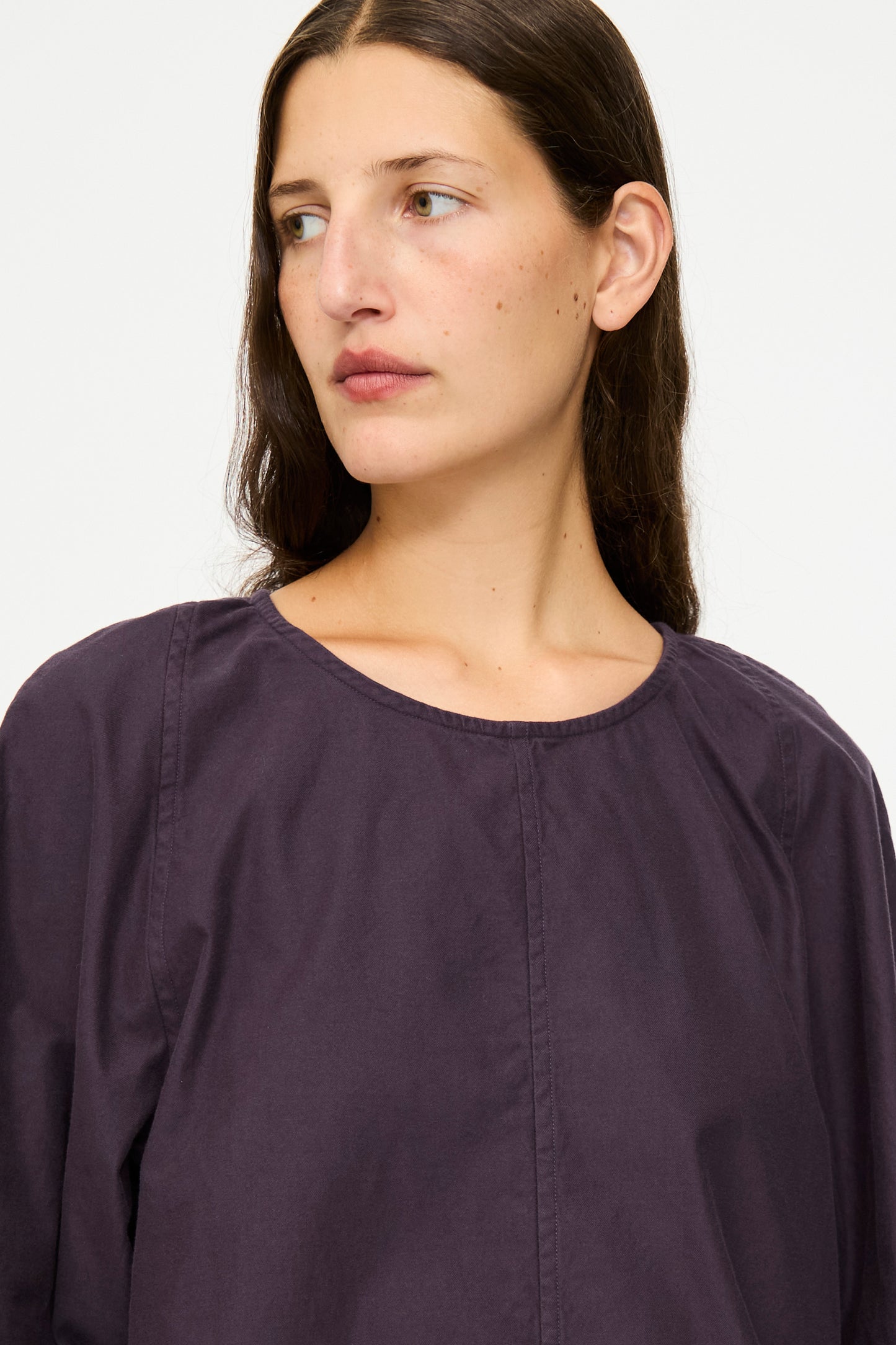 Woman with long brown hair wears the SONO Tiga Top in Dark Grape, an oversized cotton poplin shirt, while looking slightly to the side against a plain background.