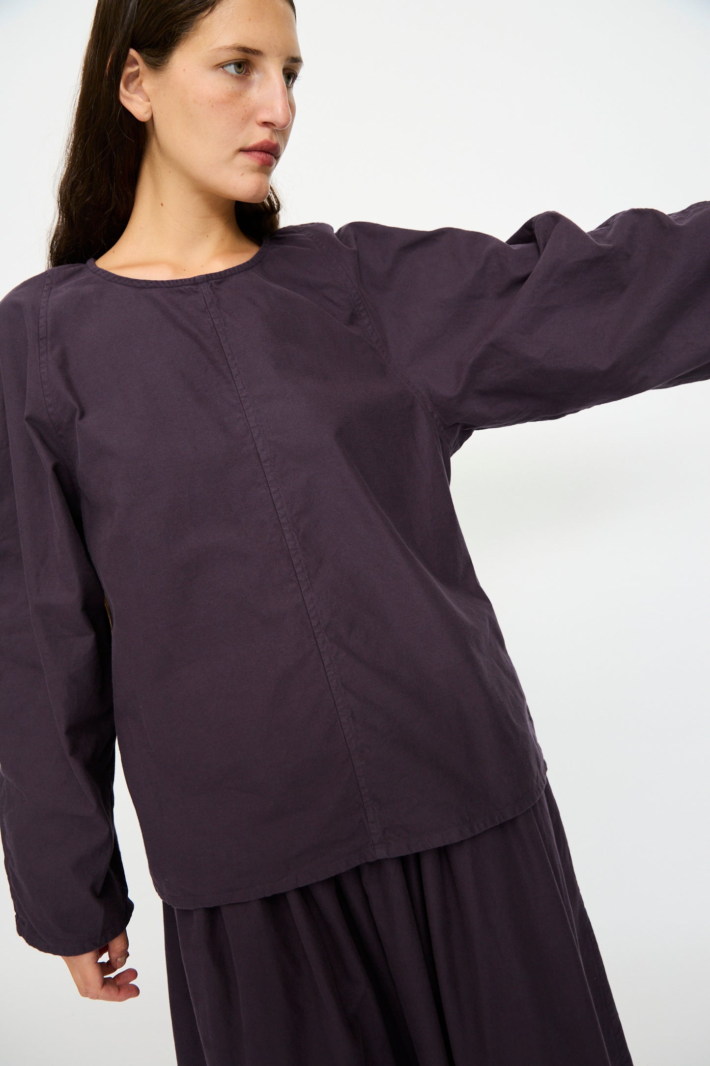 A person with long brown hair wears the SONO Tiga Top in Dark Grape, an oversized, long-sleeve organic cotton shirt, standing against a plain white background with one arm extended.
