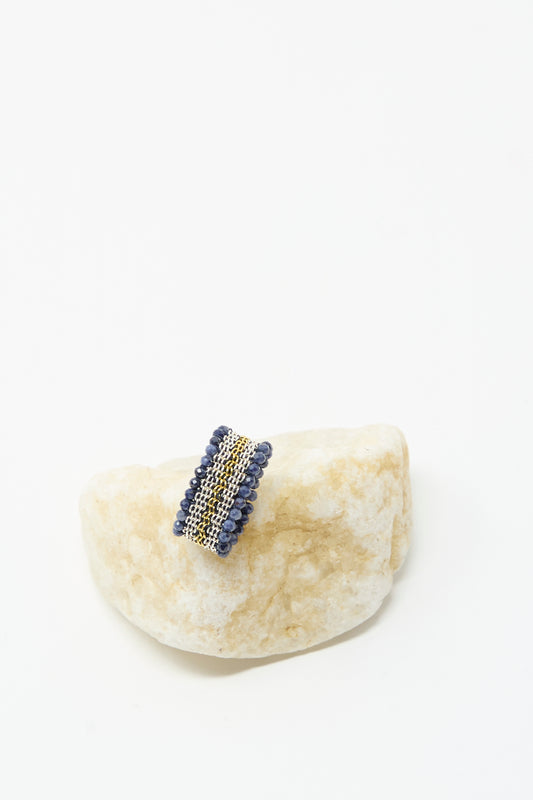 The Stephanie Schneider Silver, Gold Plated and Silk Woven Ring with Blue Sapphires is displayed on a light stone against a white background.