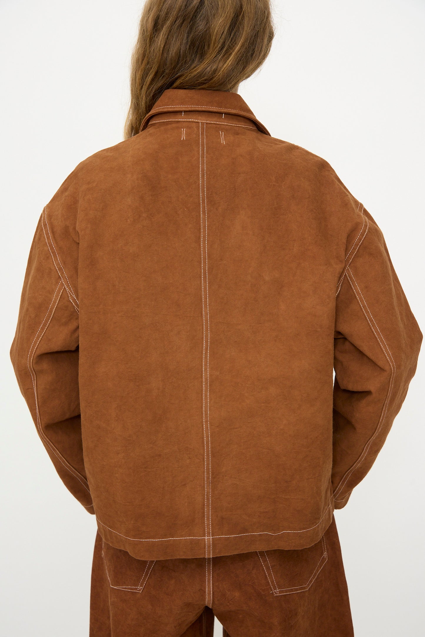 A person with long brown hair wears the Story mfg. Canvas Cove Jacket in Brown Wonky-Wear, paired with matching pants, photographed from the back against a plain light background.