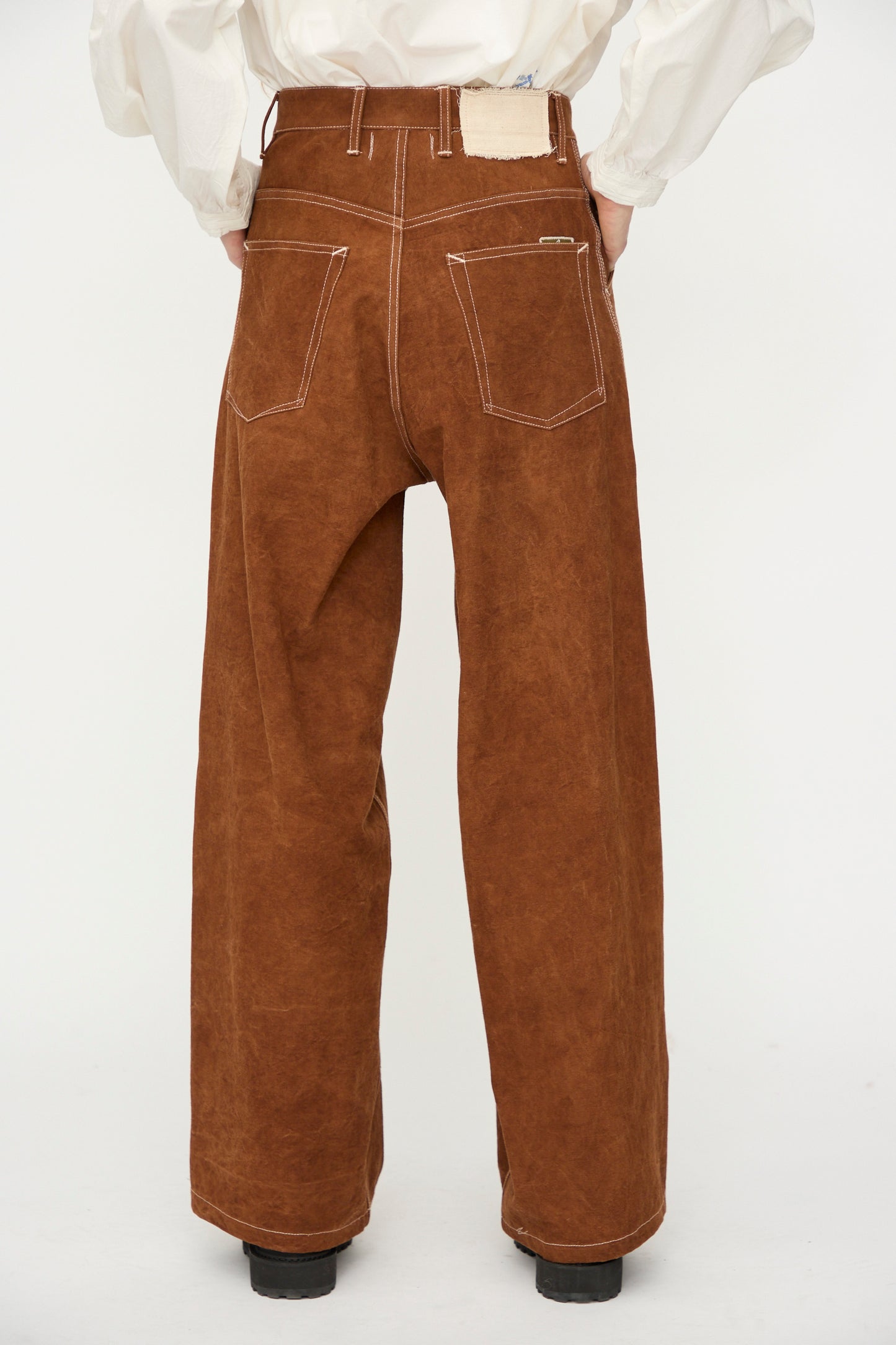 Person wearing Story mfg.'s Canvas Home Pant in Brown Wonky-Wear—organic cotton, high-waisted, wide-leg with white stitching. Back view; hands in back pockets against a plain white background.